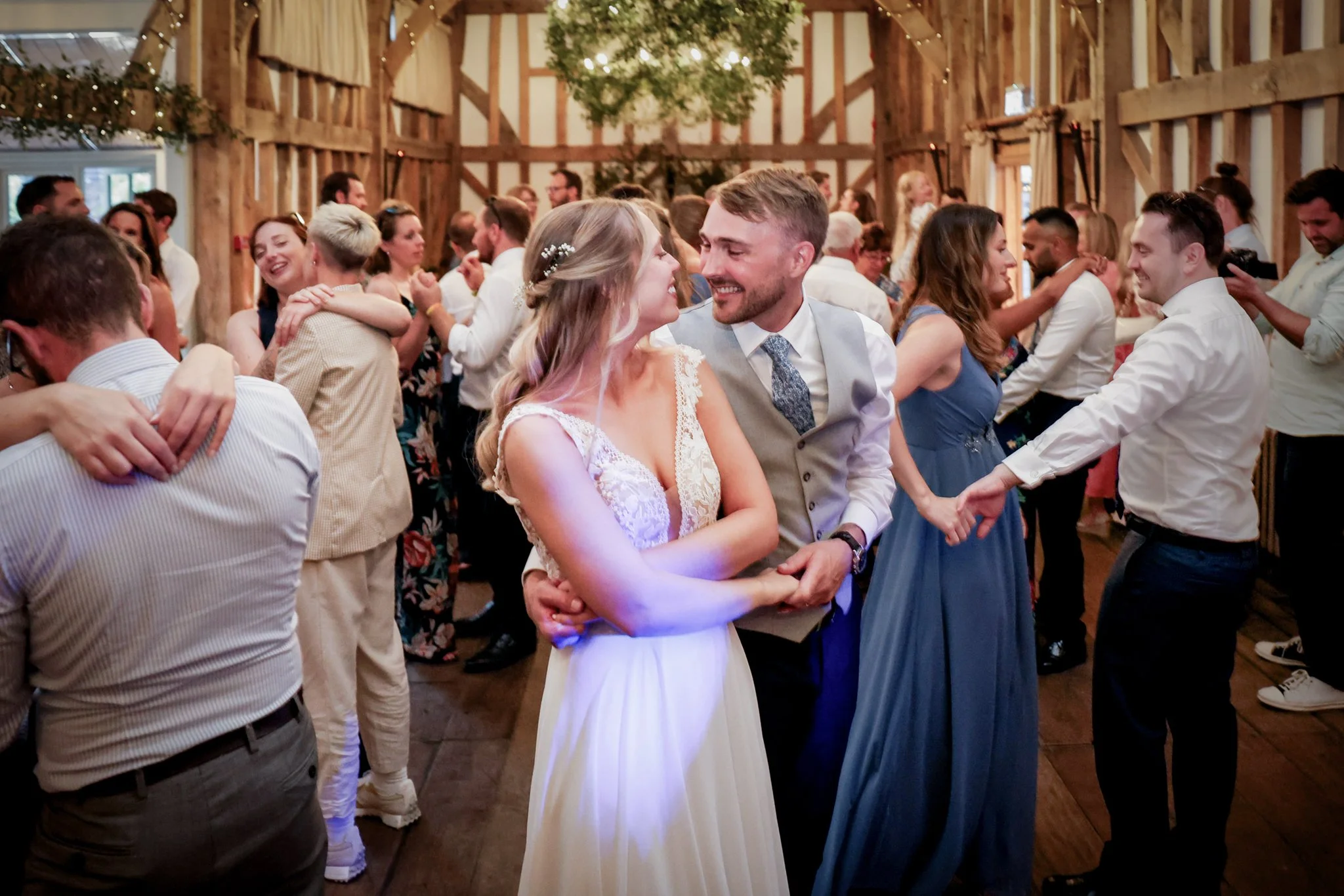 A couple dancing closely at a wedding reception, surrounded by other dancing guests in a rustic wooden venue decorated with string lights and greenery.