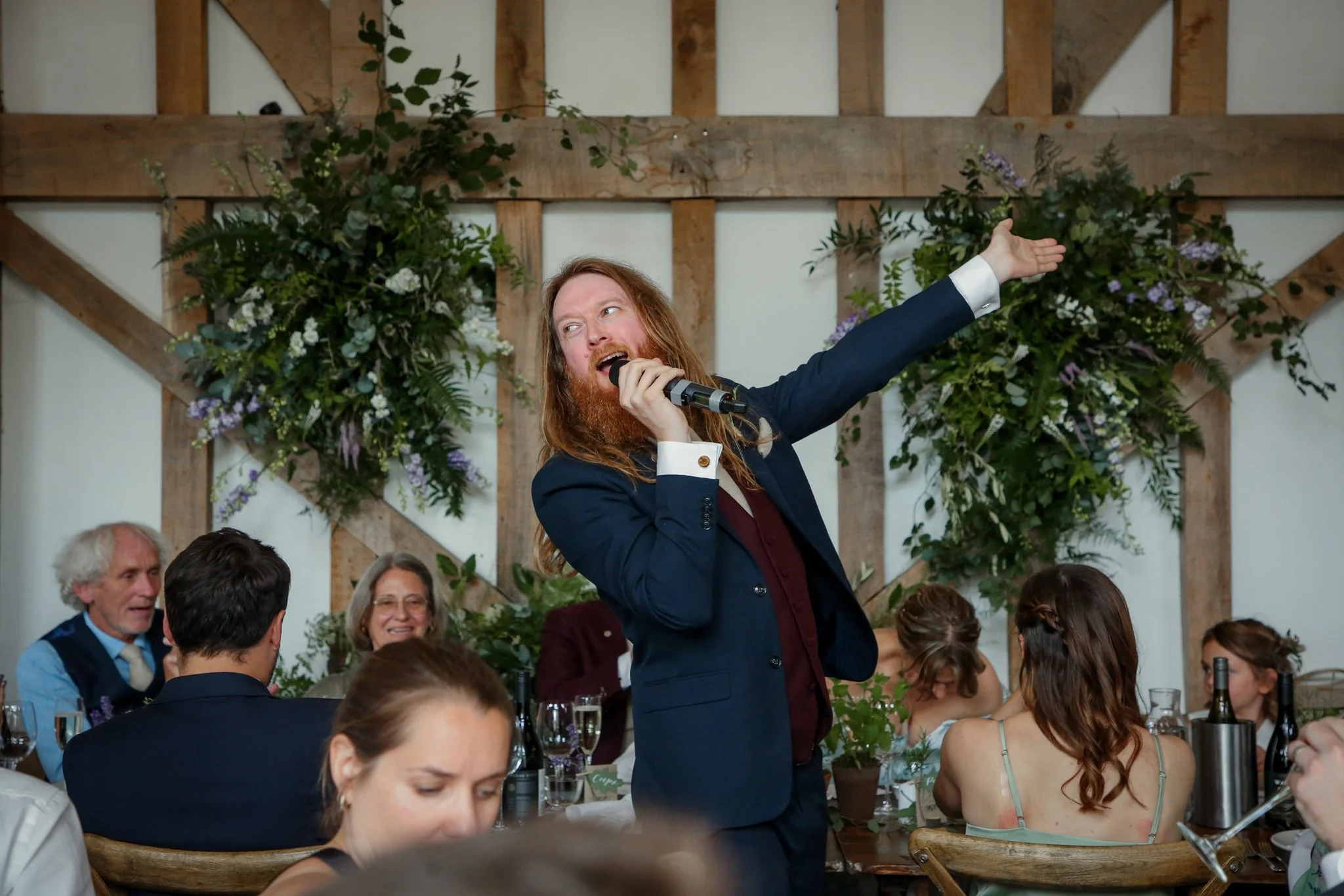 A man with long hair and a beard, dressed in a suit, is singing or speaking into a microphone while standing at a social gathering with people seated at tables decorated with flowers. The venue has wooden beams and floral decorations on the wall.