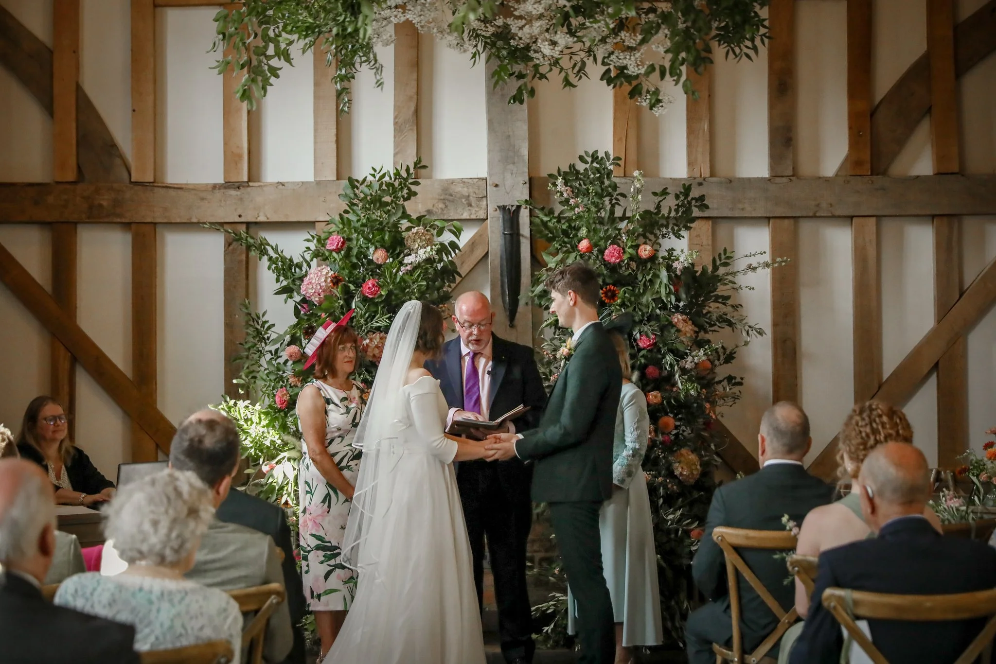 A wedding ceremony with a bride and groom exchanging vows in front of an officiant, surrounded by guests, floral arrangements, and wooden barn-like interior.