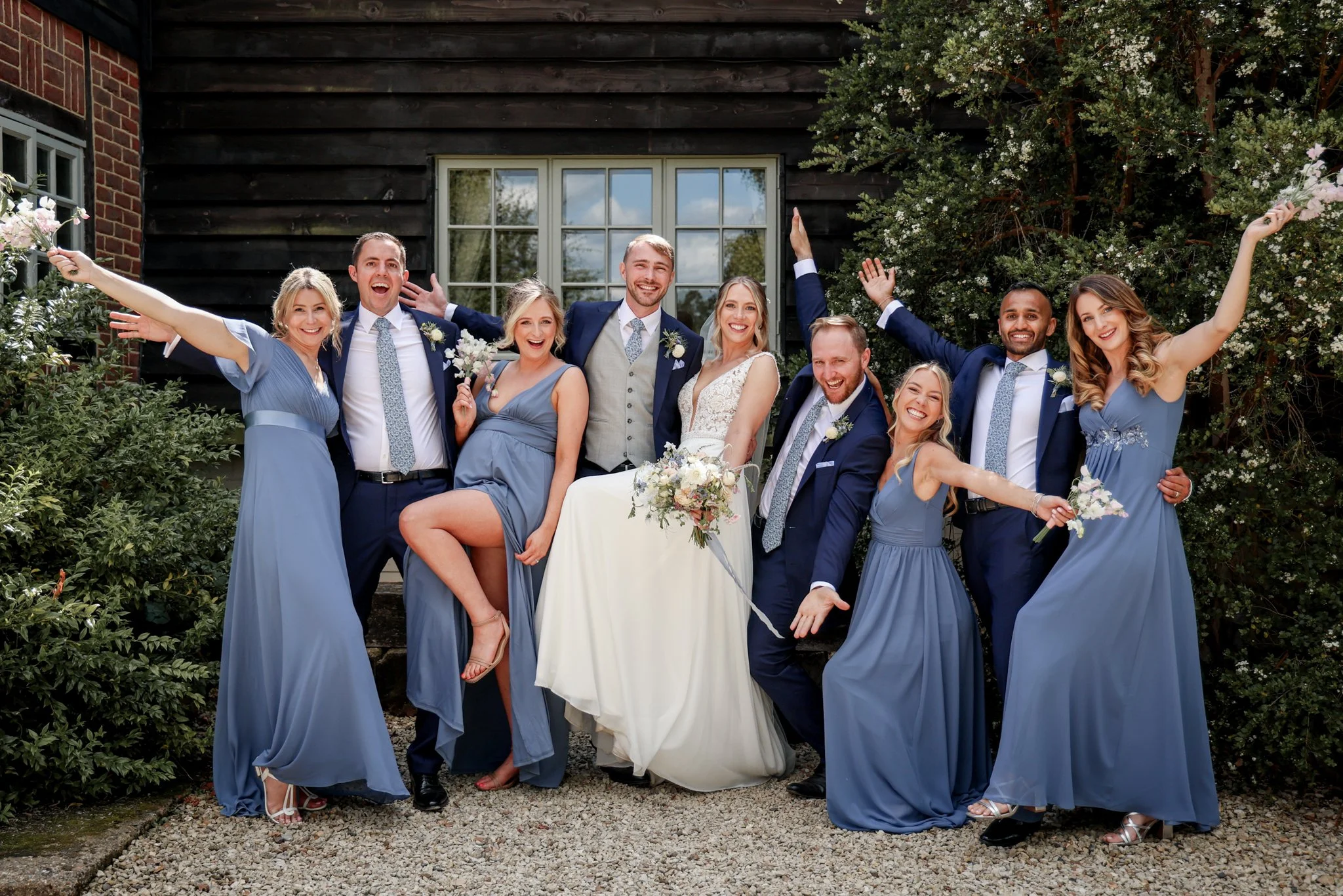 A group of people celebrating at a wedding, with women in blue dresses and men in suits, standing outdoors in front of a building with a window and greenery.