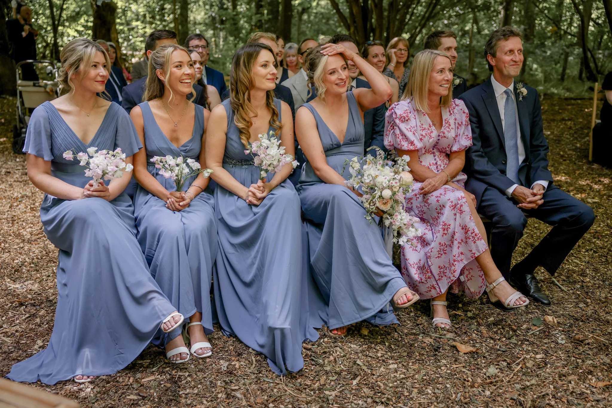 A group of women in blue dresses and a woman in a pink dress sitting outdoors in a forest, attending a wedding or special event, with smiling and happy expressions.