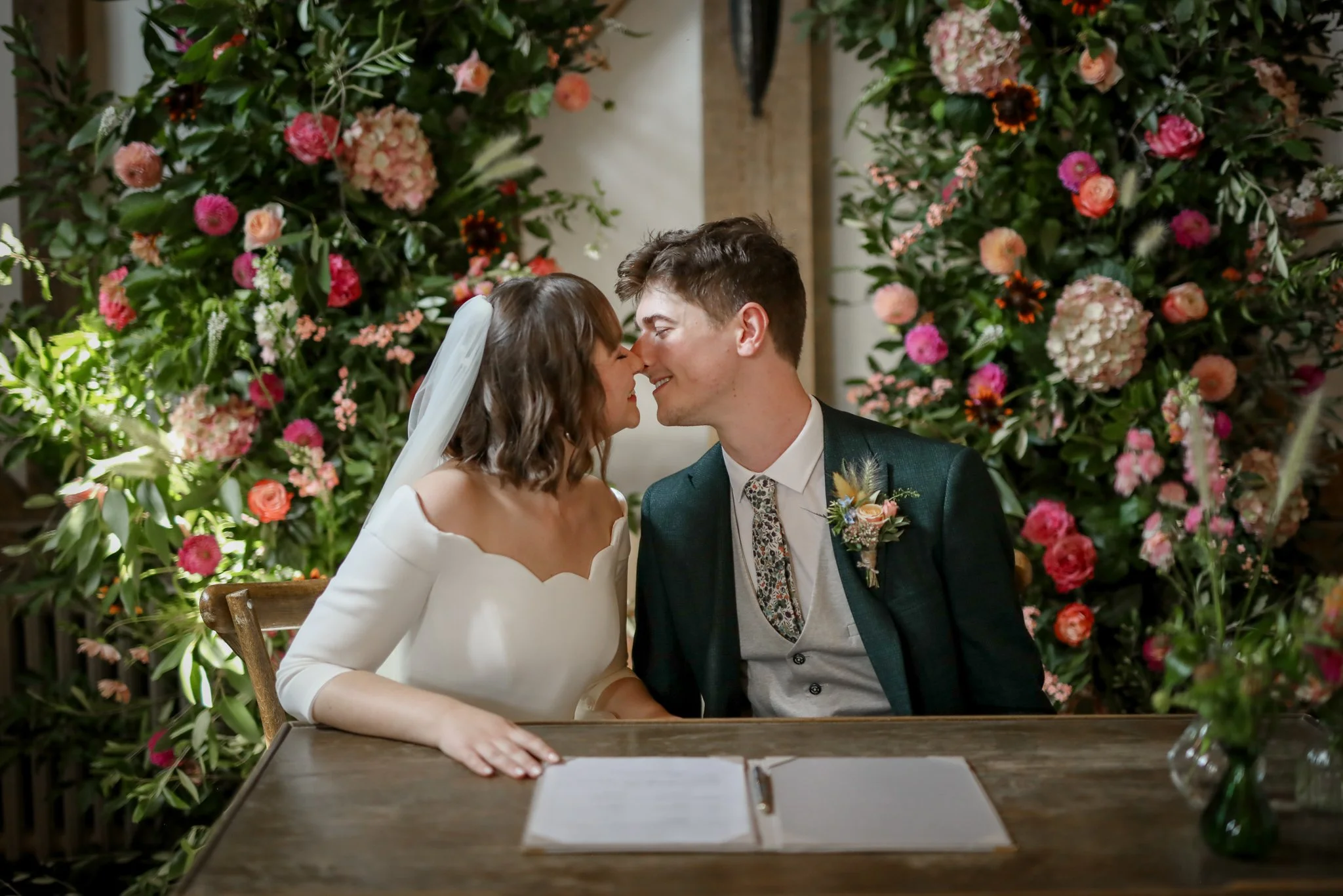 A bride and groom sitting close together at their wedding ceremony, touching noses and smiling, with a background of pink and white flowers.