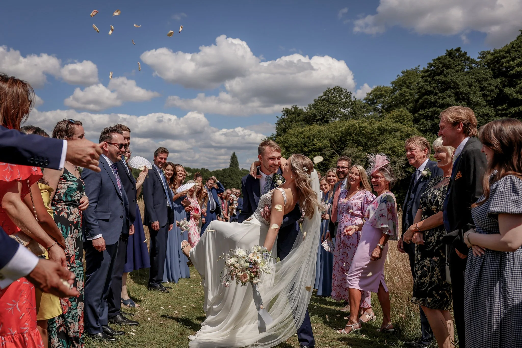 A wedding ceremony outdoors with guests smiling and looking at a bride and groom who are hugging, with floating rose petals in the air under a partly cloudy sky.