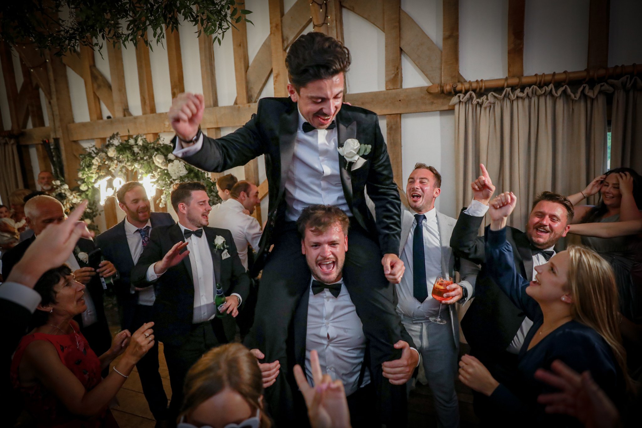 A man in a tuxedo is being lifted and celebrated by a group of people at a wedding reception. Everyone appears to be happy and cheering, with some holding drinks.