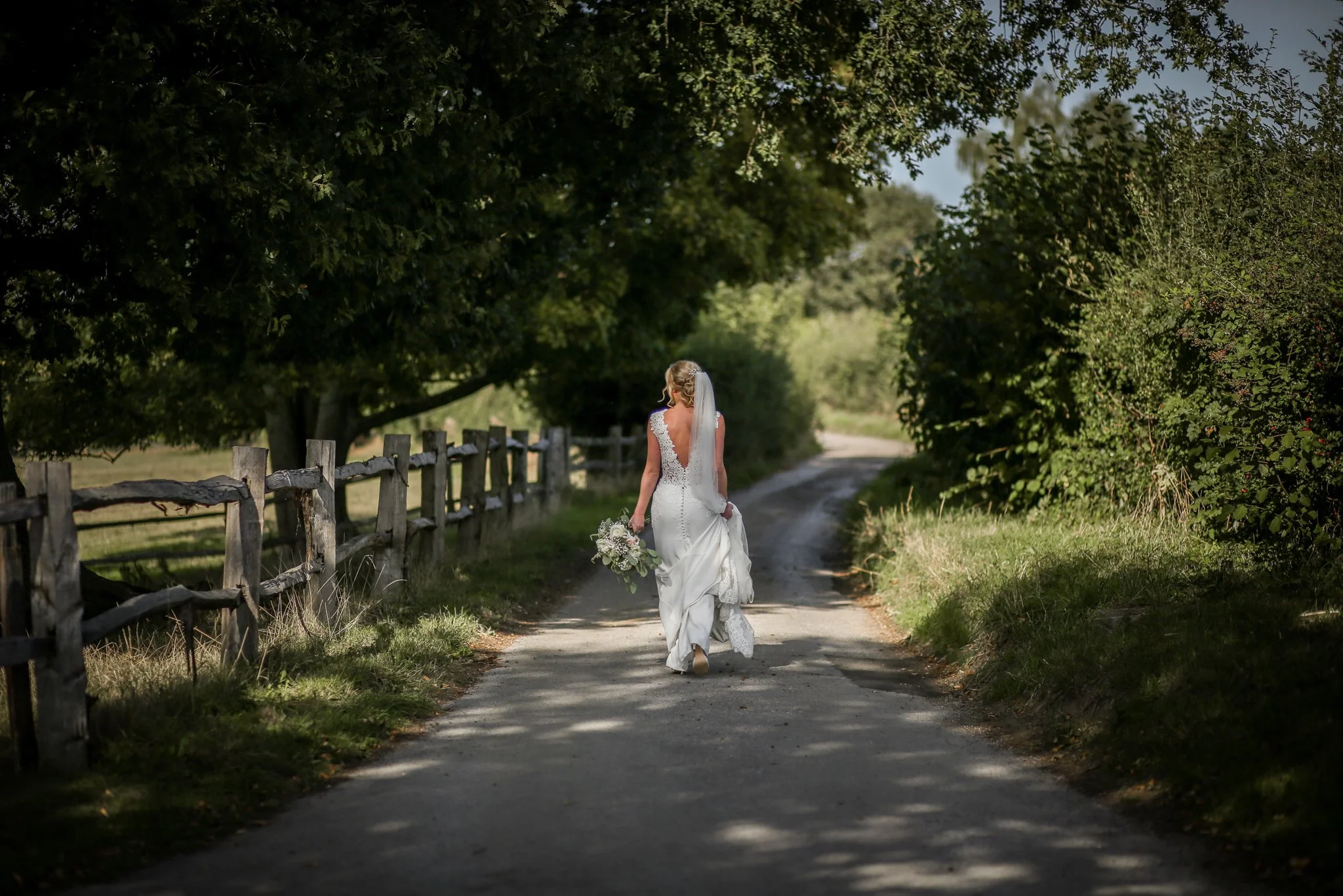 A bride walking along a dirt road lined with wooden fences and lush green trees, carrying a bouquet, wearing a white lace wedding dress and veil.