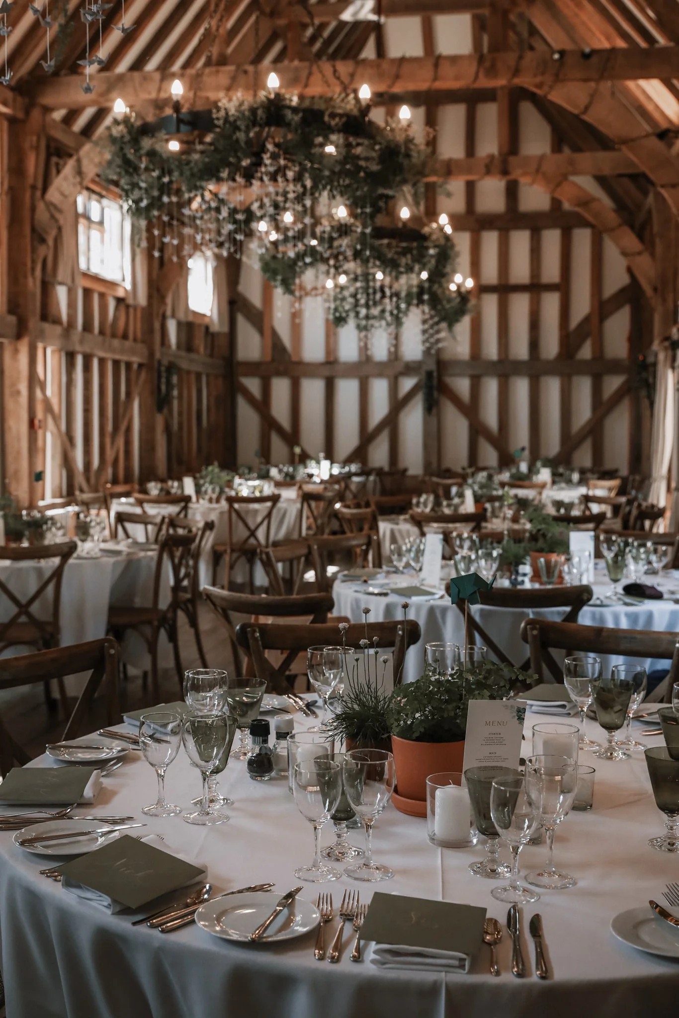 Interior of a rustic barn decorated for a wedding or celebration with round tables, white tablecloths, glassware, silverware, and a small potted plant centerpiece. The barn features wooden beams and chandeliers with greenery and lights hanging from t
