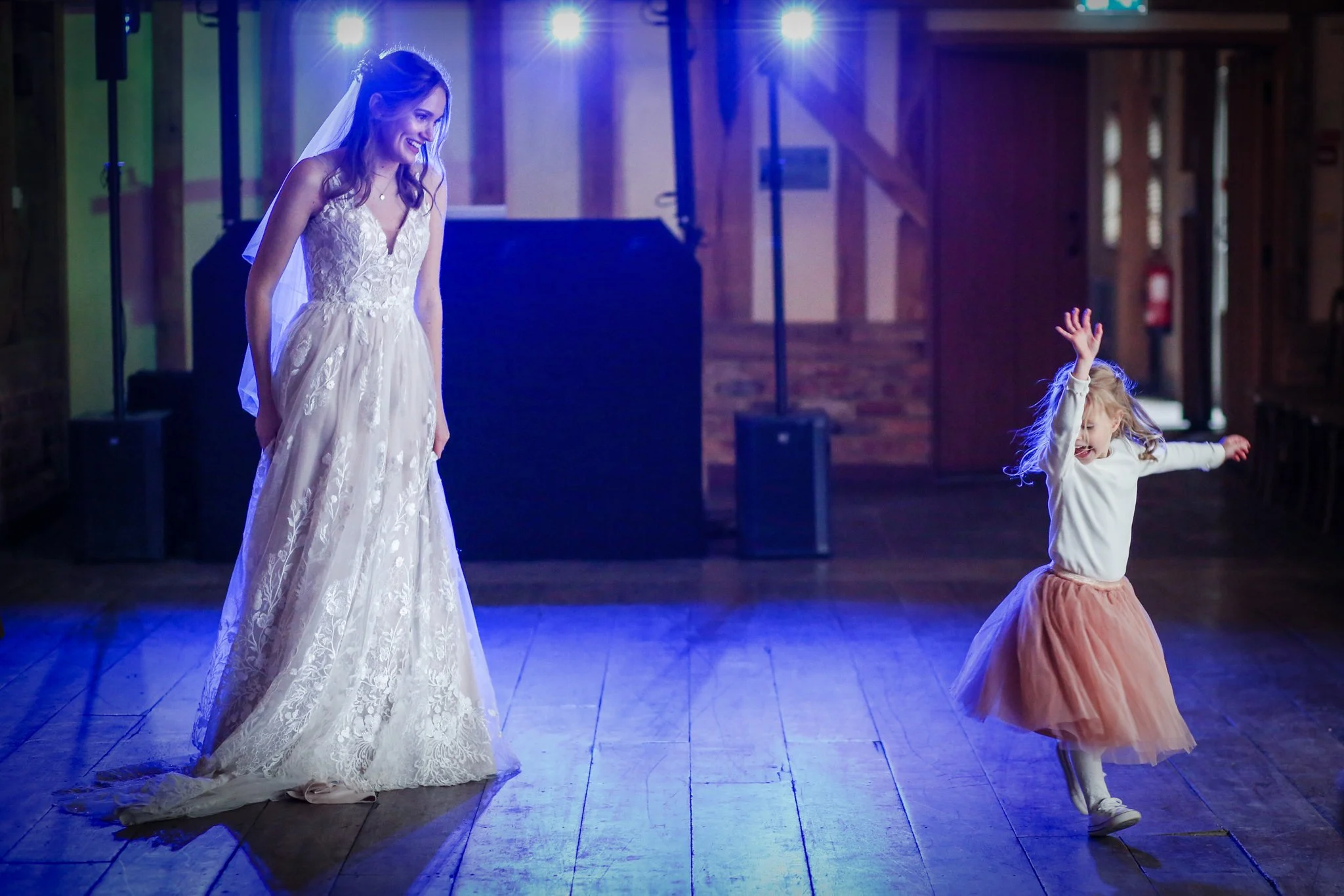 A woman in a wedding dress is smiling and looking down at a young girl in a tulle skirt who is joyful, raising her hand, and spinning in a dance. They are in a dimly lit room with blue stage lighting.