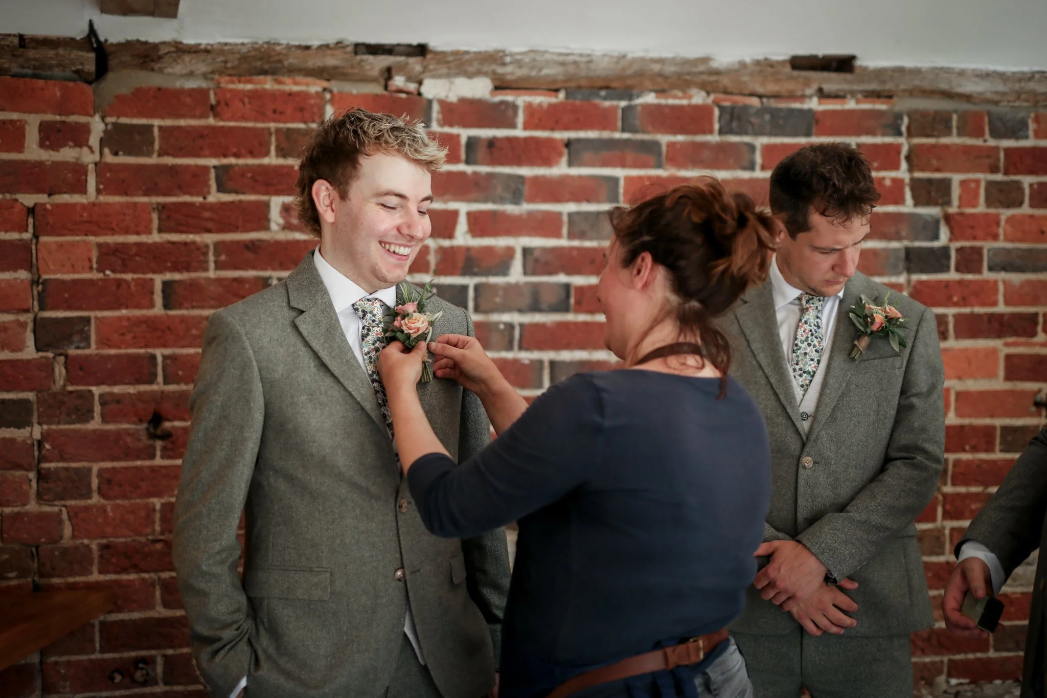 A woman pinning a boutonniere on a young man in a gray suit during a wedding ceremony, with another man in a suit standing nearby against a brick wall.
