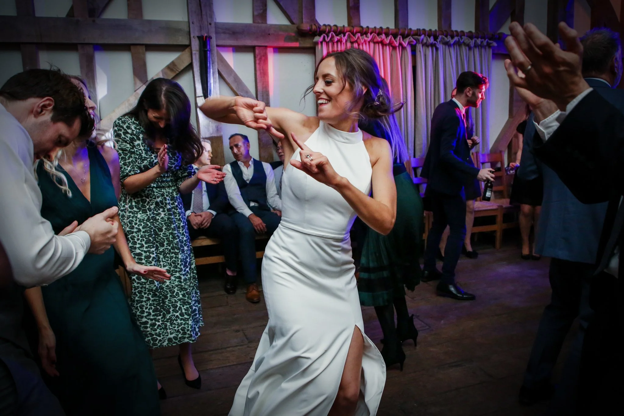 A woman in a white dress dancing and smiling at a lively party with other guests dancing and socializing in the background.