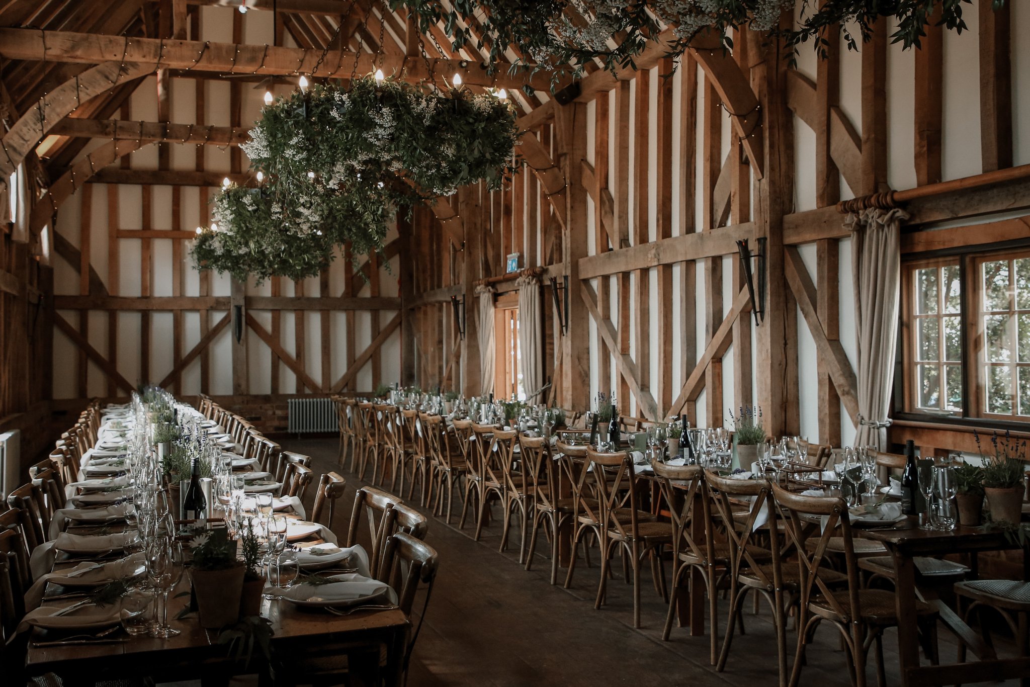 Interior of a rustic wedding or event venue with wooden beams, long tables set with white tablecloths, wine bottles, glassware, and floral centerpieces, and a large hanging greenery arrangement with lights.
