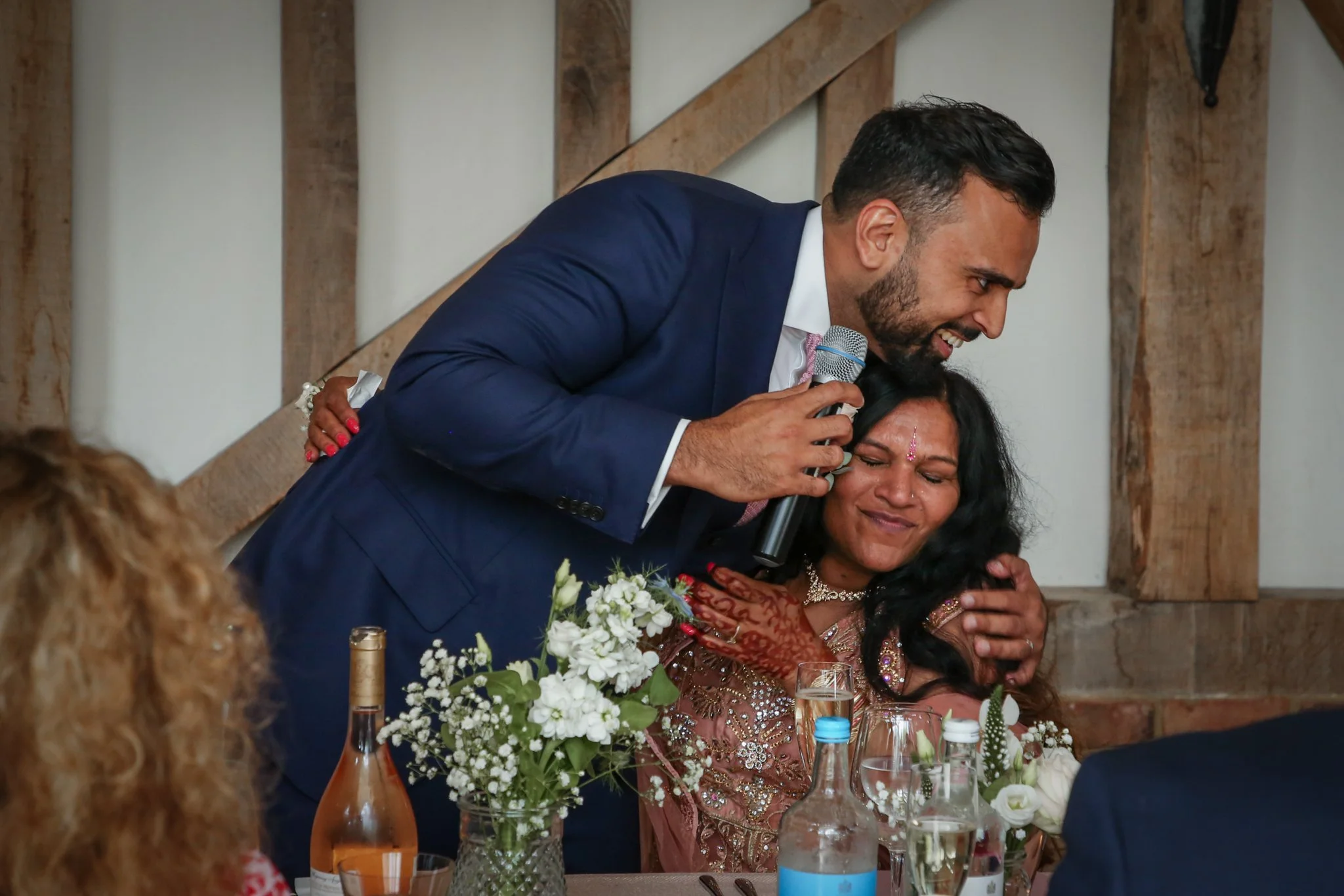 A man in a blue suit is leaning over and embracing a woman dressed in traditional Indian attire at a celebration, likely a wedding reception. The man is holding a microphone and smiling. The woman is seated at a table decorated with white flowers and