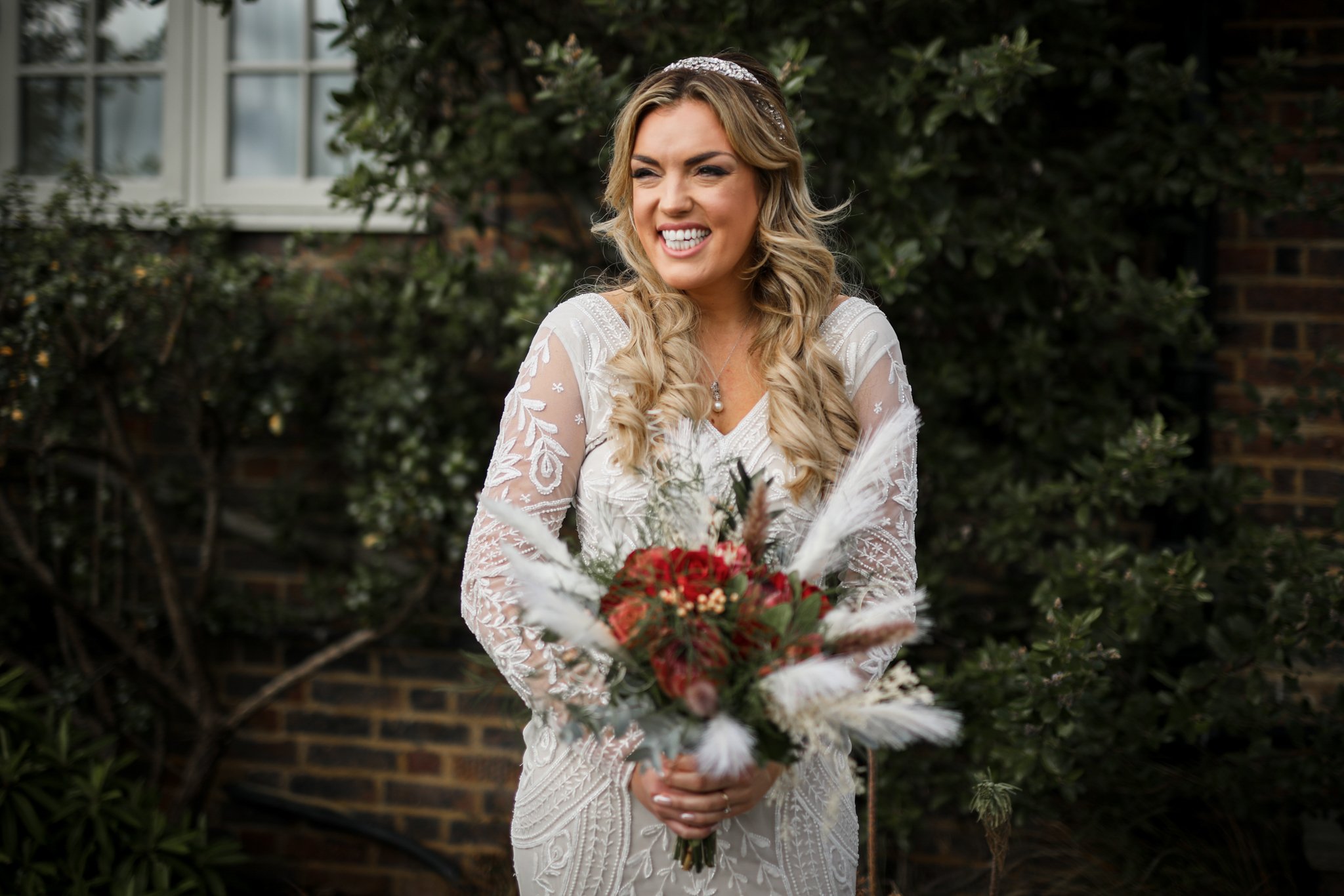 A woman in a white lace wedding dress holding a bouquet of flowers, smiling and looking to her right, outdoors with green foliage and a brick wall in the background.
