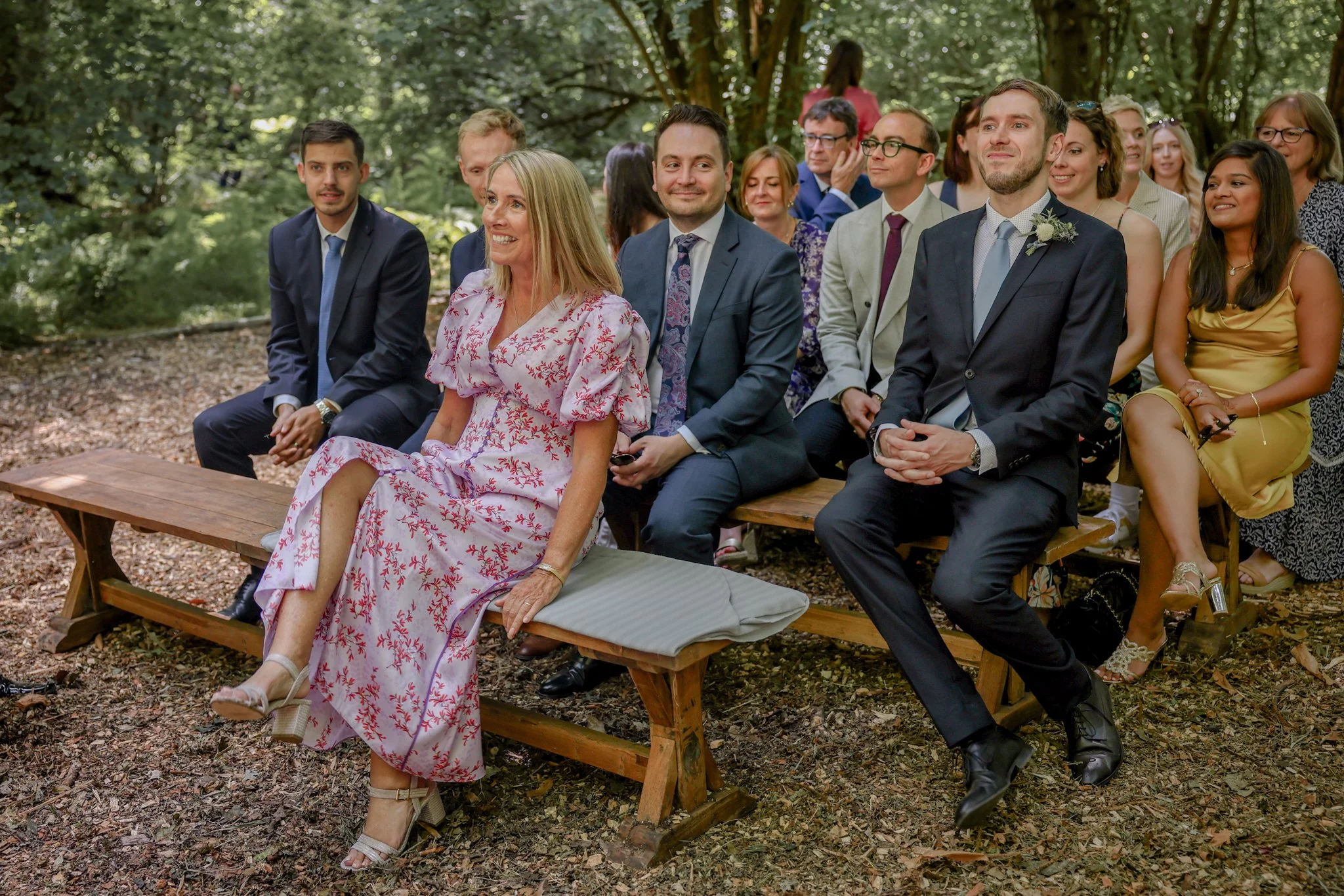 A group of men and women dressed in formal attire, sitting on wooden benches outdoors among trees, attending a wedding or similar event.