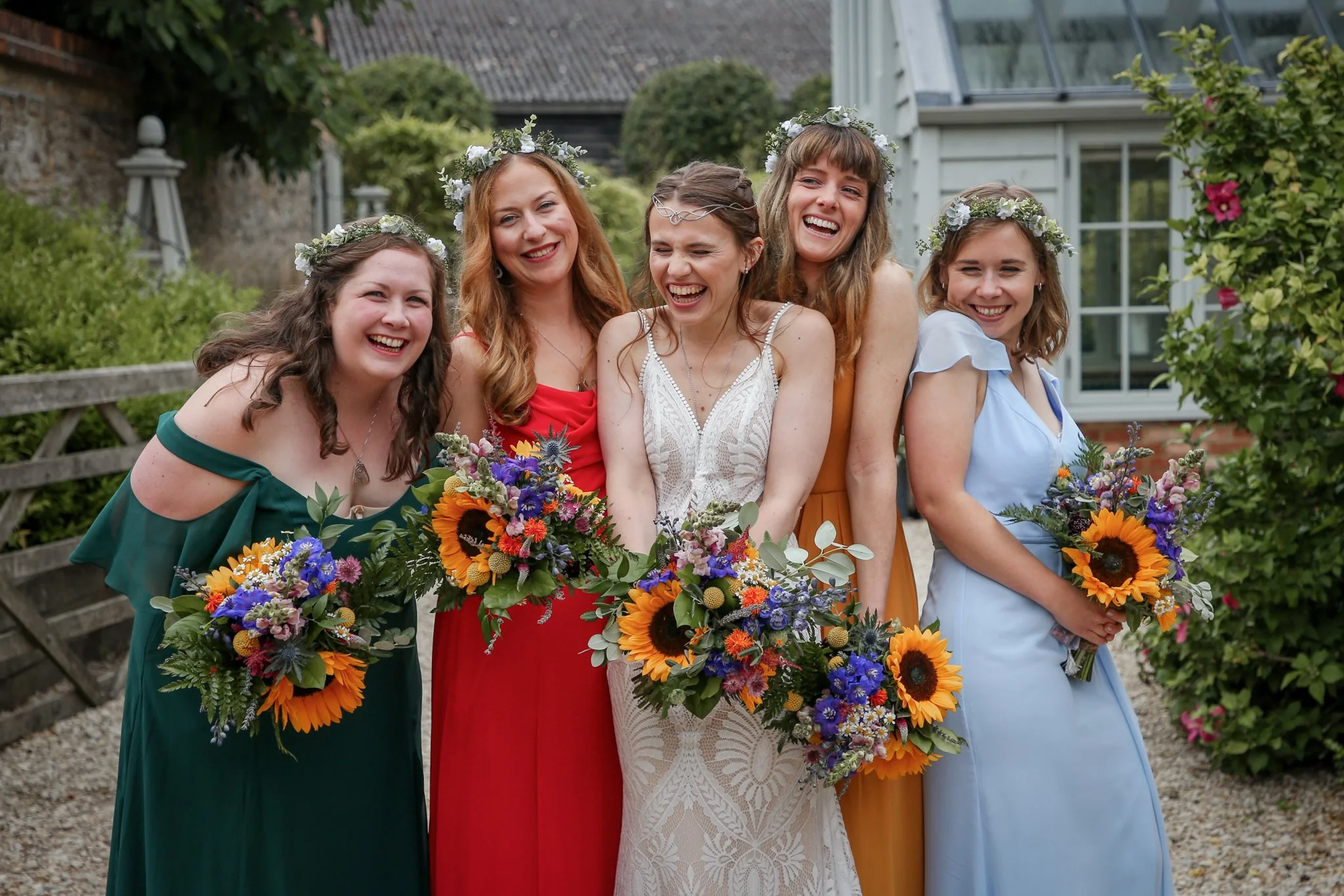 Group of five women wearing colorful dresses, floral crowns, and holding bouquets, standing outdoors in a garden setting, smiling and enjoying a celebration.