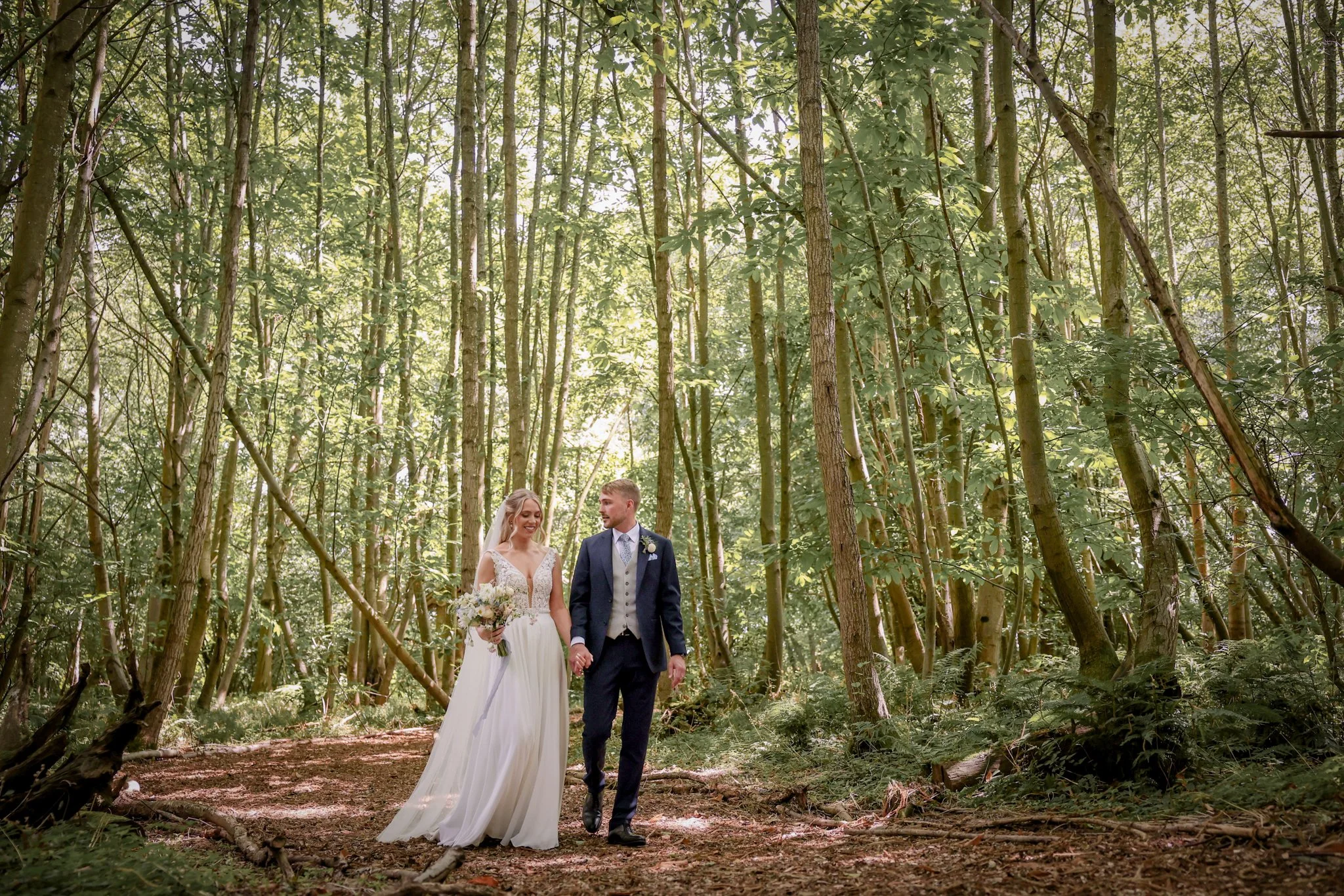 A bride and groom walking hand in hand through a forest with tall trees, sunlight filtering through the leaves, during a wedding ceremony.