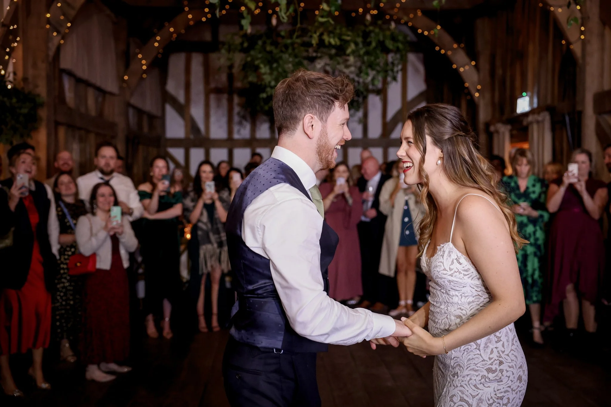 A bride and groom smiling and holding hands during their wedding dance in a rustic barn decorated with string lights, with guests watching and taking photos in the background.
