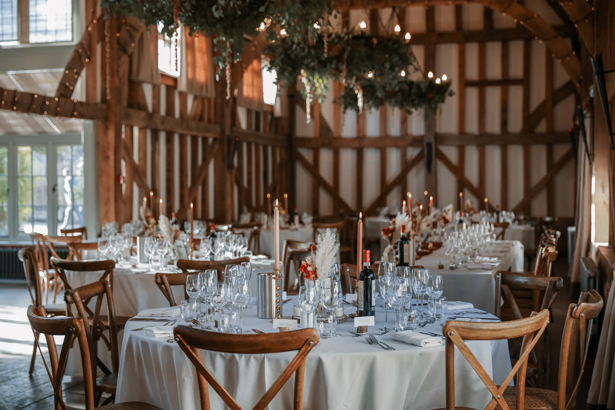 Interior of a rustic barn decorated for a celebration with long dining tables, elegant glassware, candles, and floral centerpieces, with wood beams and fairy lights overhead.