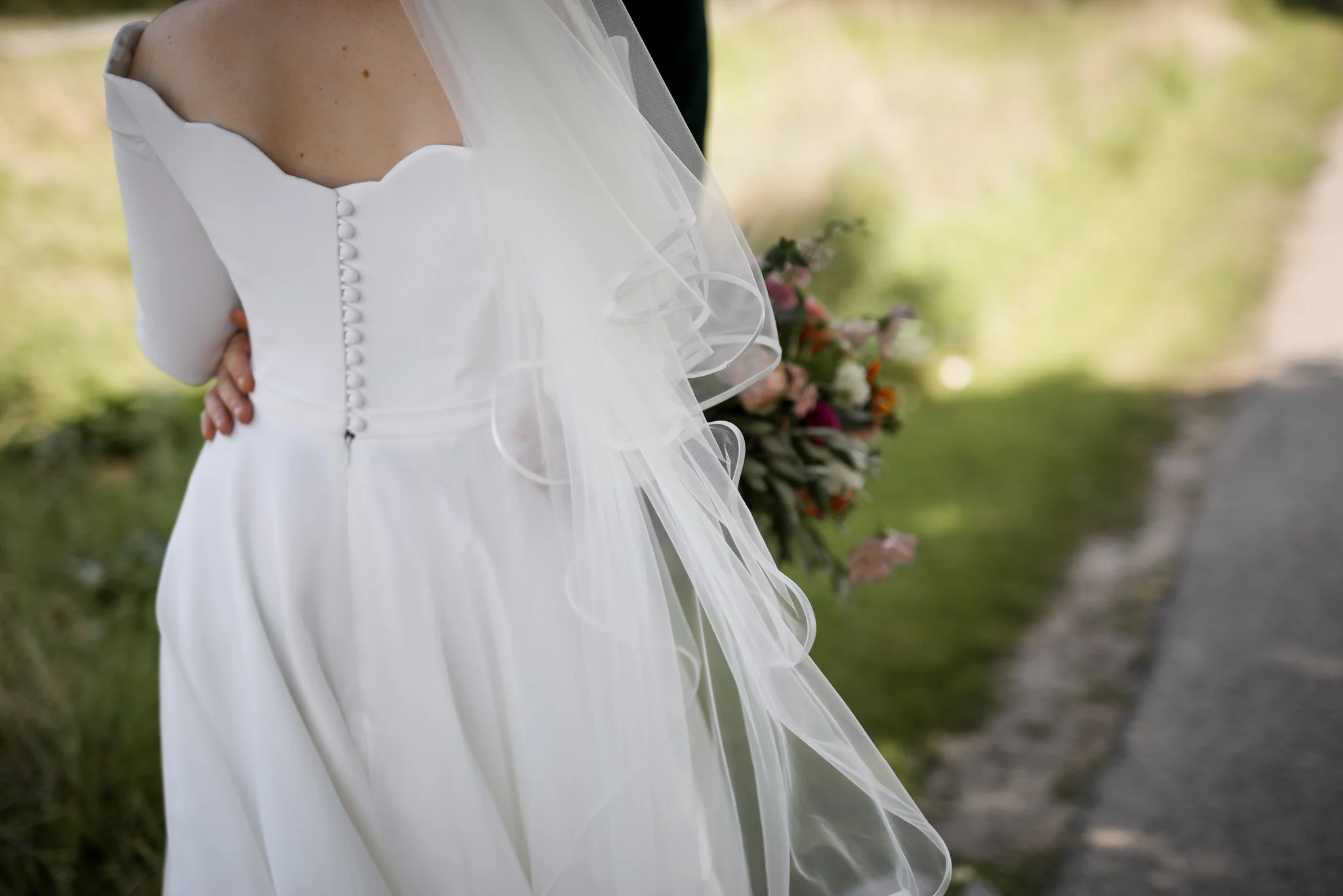 Close-up of a bride in a white wedding dress with buttons on the back, holding hands with a groom, outdoors with green trees and a pathway in the background.