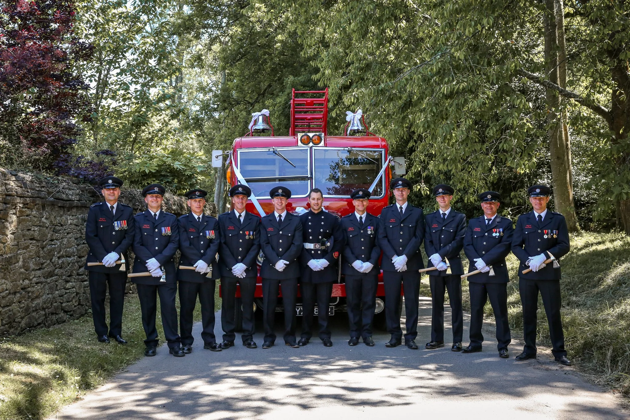 Group of ten firefighters in uniform standing in front of a red fire truck outdoors surrounded by trees.