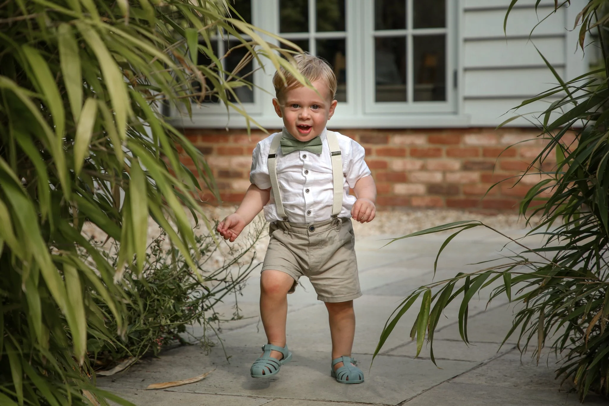 Young boy wearing a green bow tie, suspenders, a white shirt, khaki shorts, and blue sandals walking outdoors between tall green plants on a patio with a house in the background.