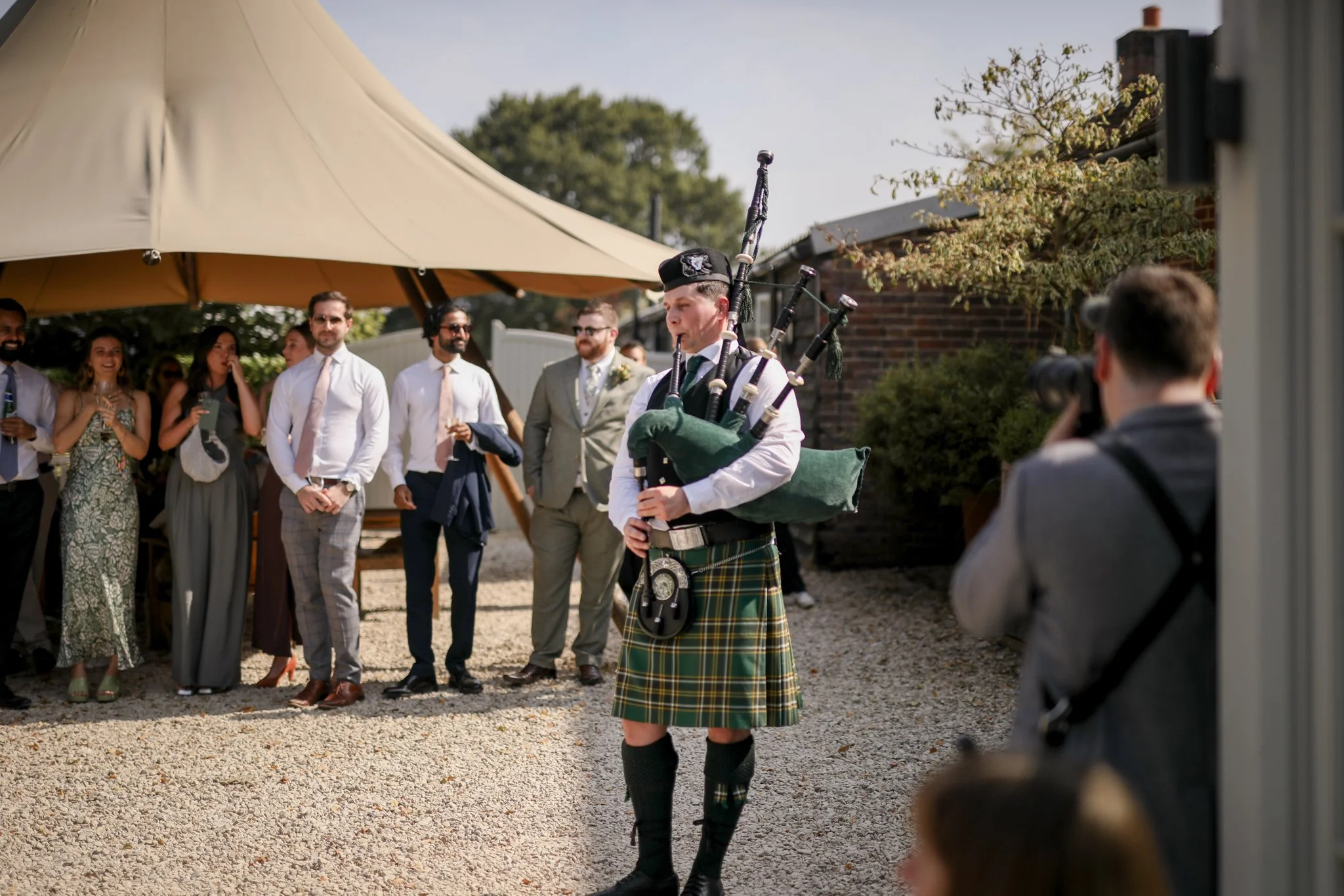 A man dressed in traditional Scottish attire, playing bagpipes at an outdoor wedding or celebration. Several well-dressed guests are standing under a large beige umbrella, watching and smiling.