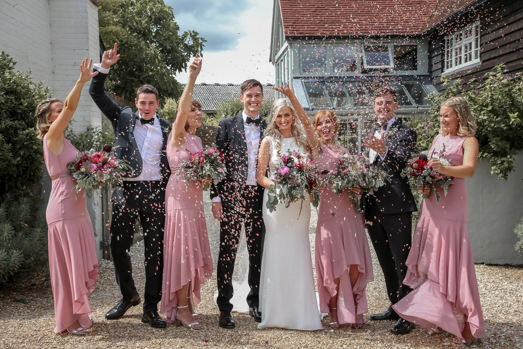 A wedding celebration outdoors with the bride, groom, bridesmaids, and groomsmen throwing pink and white confetti in the air. The bride is in a white dress, holding a bouquet, and smiling. Bridesmaids are wearing pink dresses, holding bouquets, and s