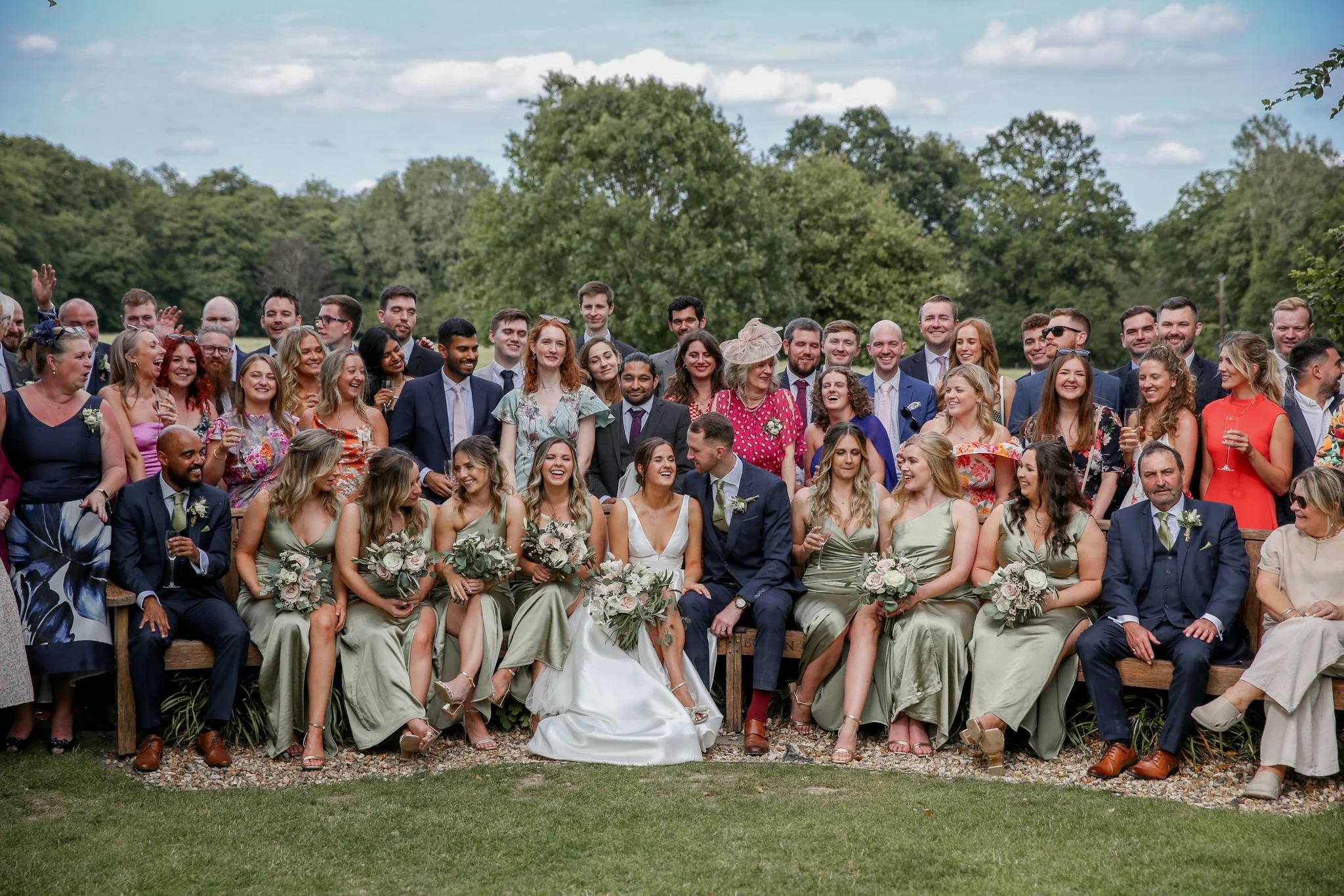 A large group of wedding guests, including men, women, and children, gathered outdoors under a partly cloudy sky, sitting and standing on wooden benches and chairs, with smiley faces and celebratory expressions.
