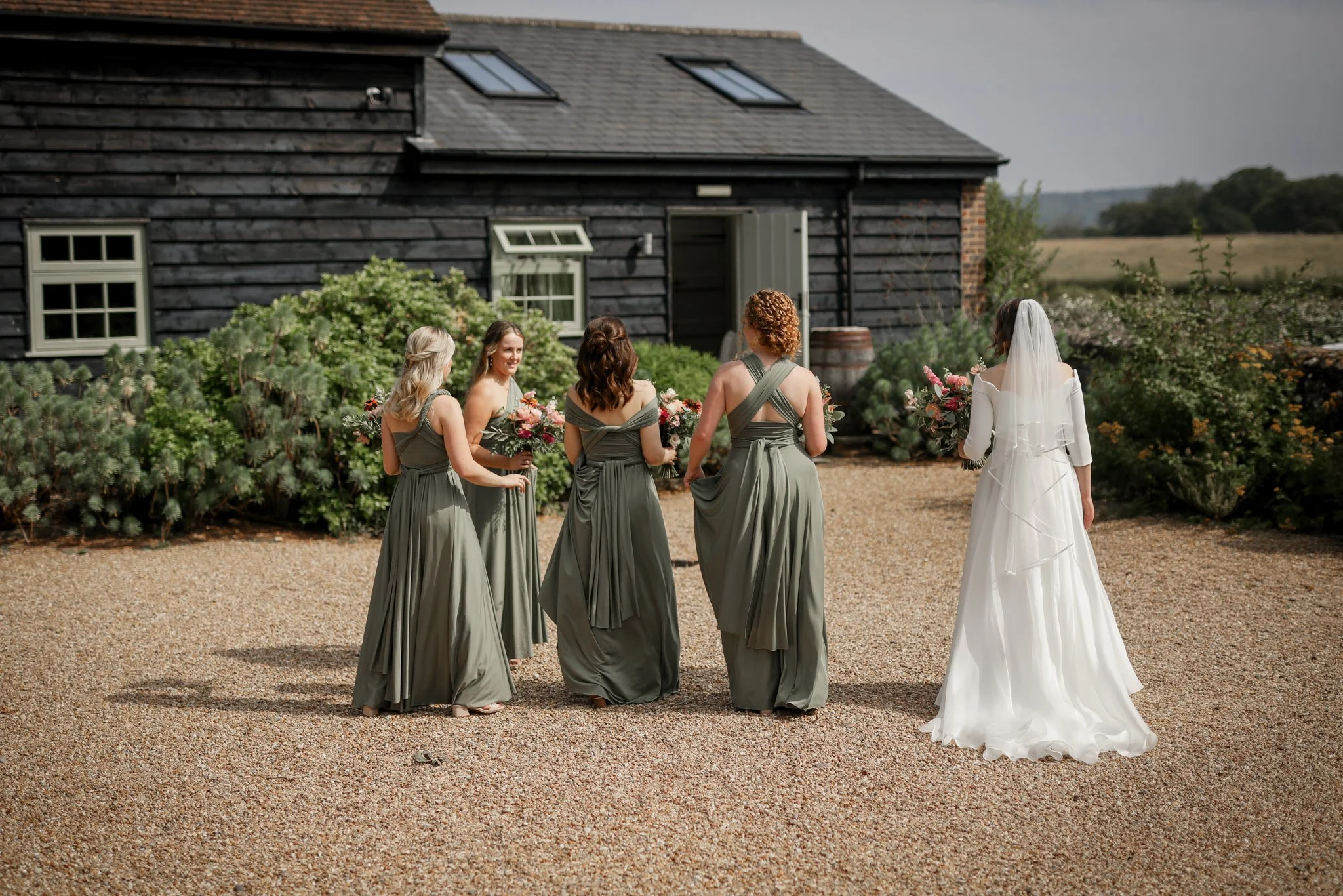 A bride in a white wedding dress with a veil stands facing away from the camera, holding a bouquet, as five bridesmaids in matching gray dresses hold their own bouquets and converse outside near a rustic black barn with a field in the background.