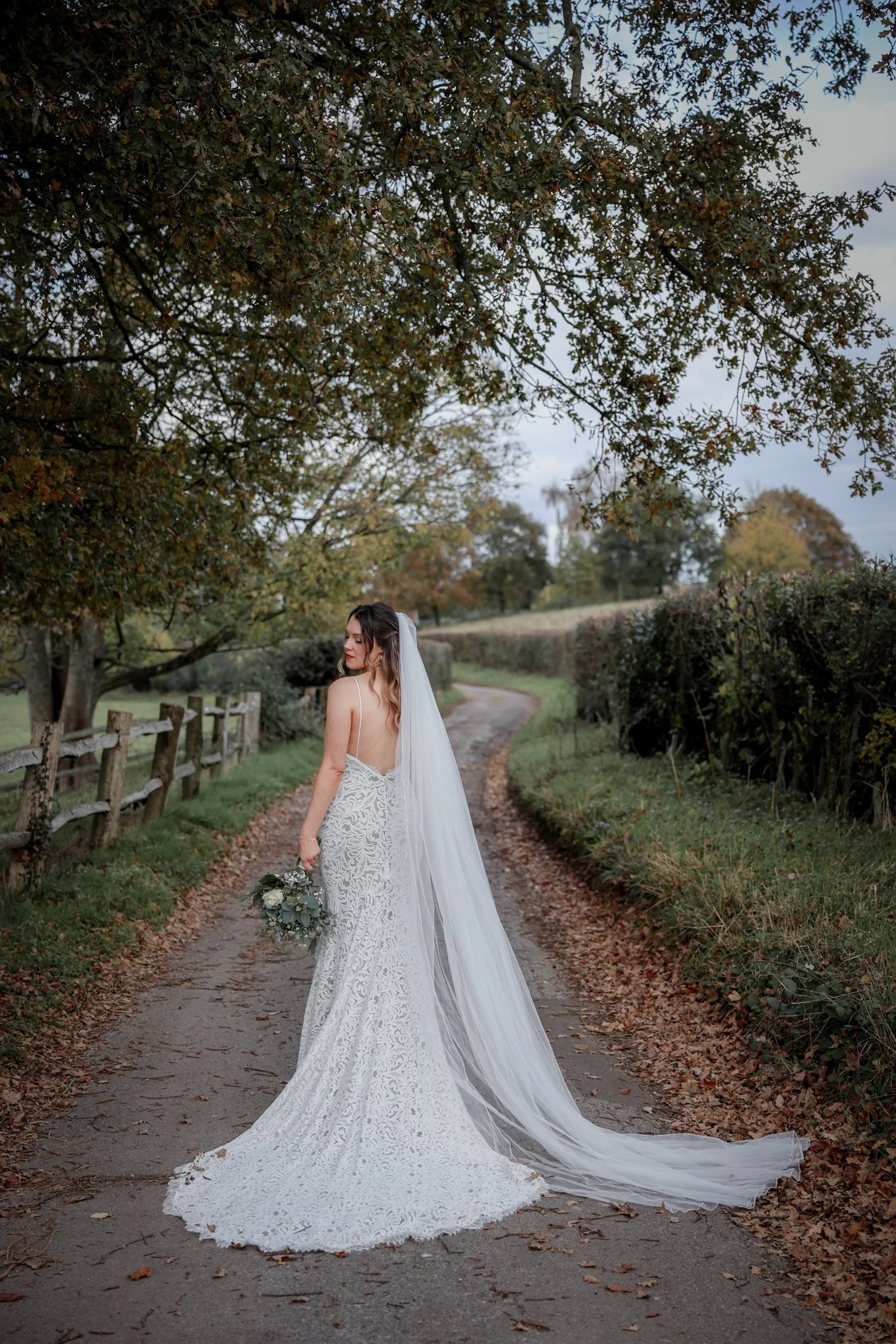 A bride in a white lace wedding gown with a long train, holding a bouquet, standing on a dirt path surrounded by trees with autumn foliage.