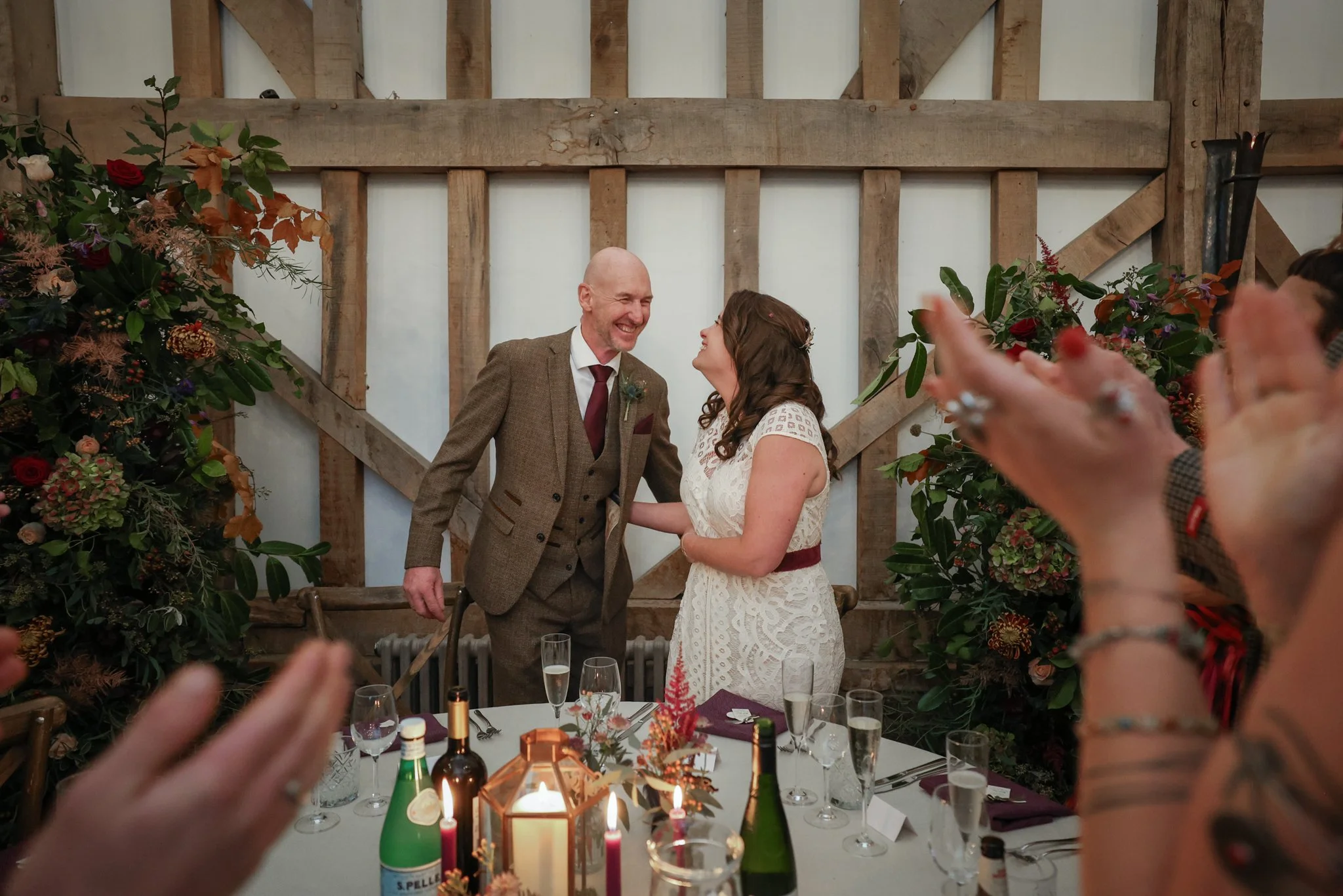 A newlywed couple standing at the head of a dinner table during their wedding reception, smiling and looking into each other's eyes, with guests clapping in the foreground, decorated with flowers and candles.