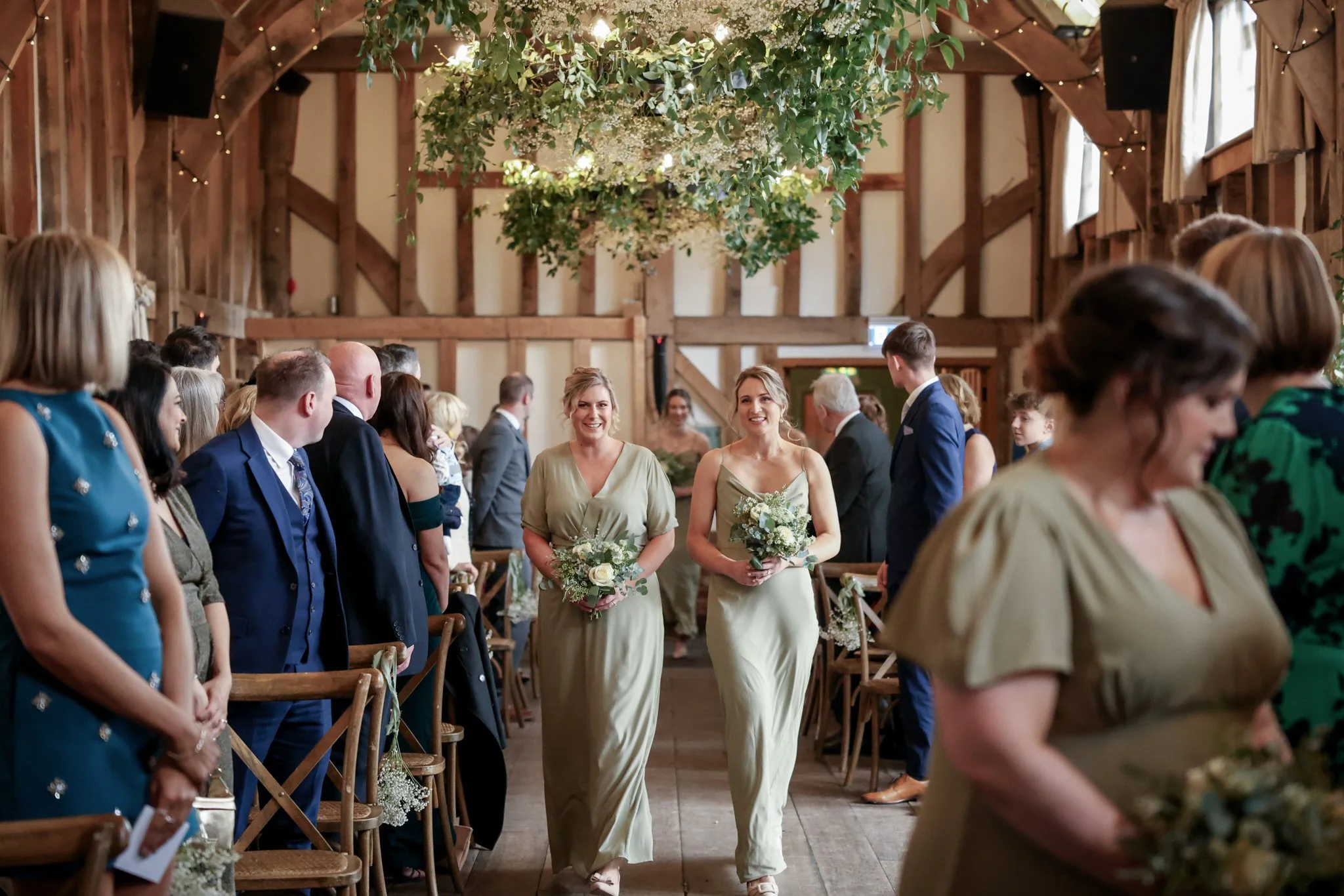Bride and bridesmaid walking down the aisle during a wedding ceremony in a rustic barn decorated with greenery and string lights, with guests seated on either side.