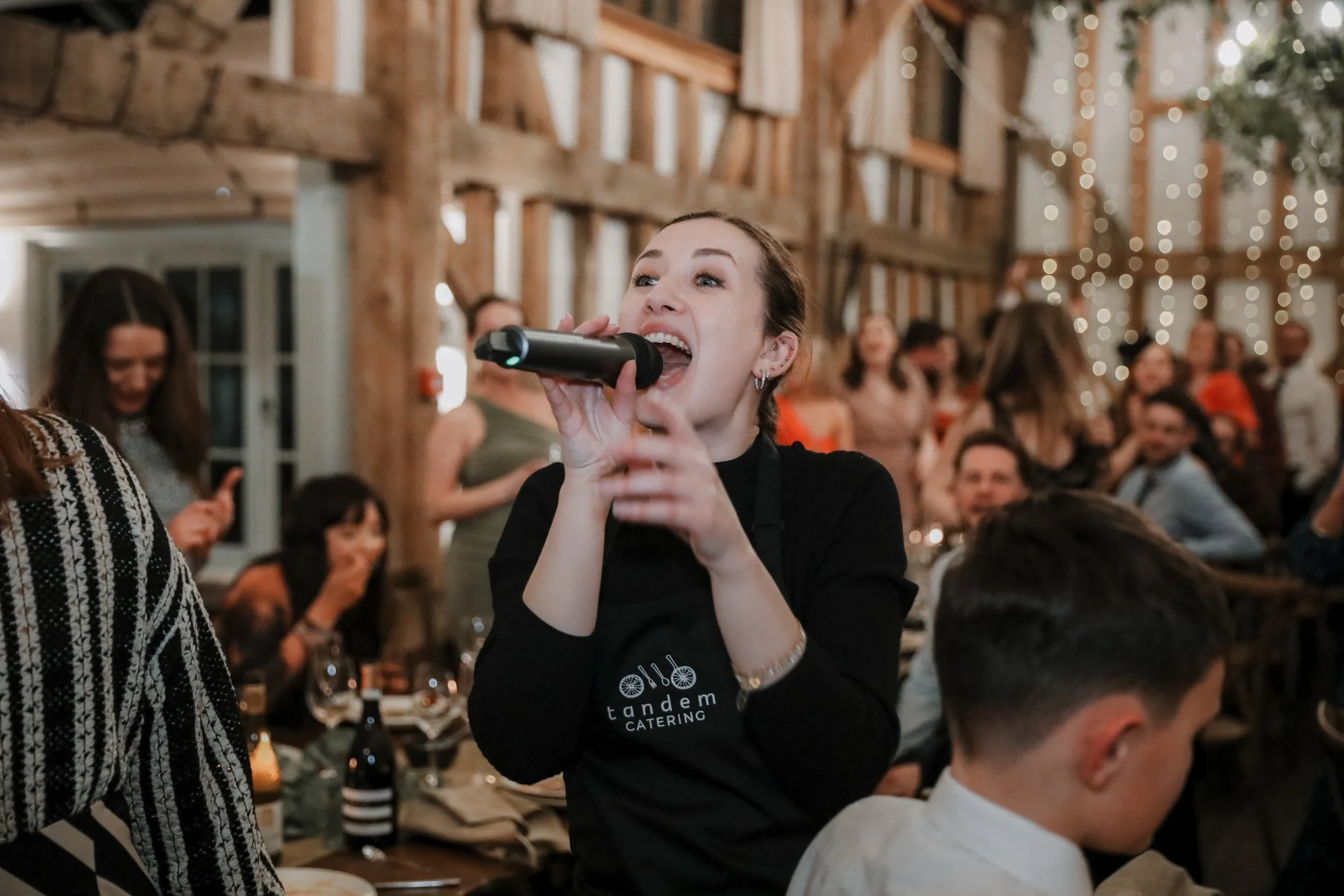 A woman singing into a microphone at a crowded indoor event with wooden walls and string lights.