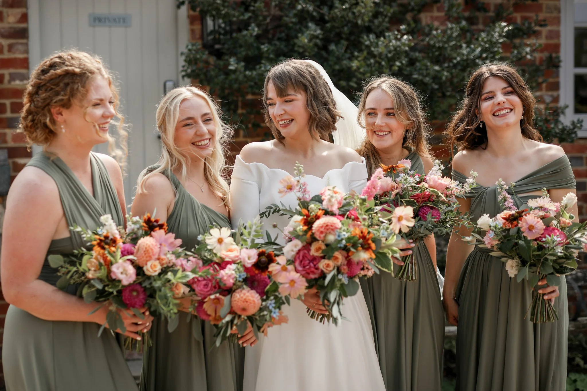 A bride in a white wedding dress standing outdoors with five women in matching sage green dresses, all holding bouquets of pink and multicolored flowers.