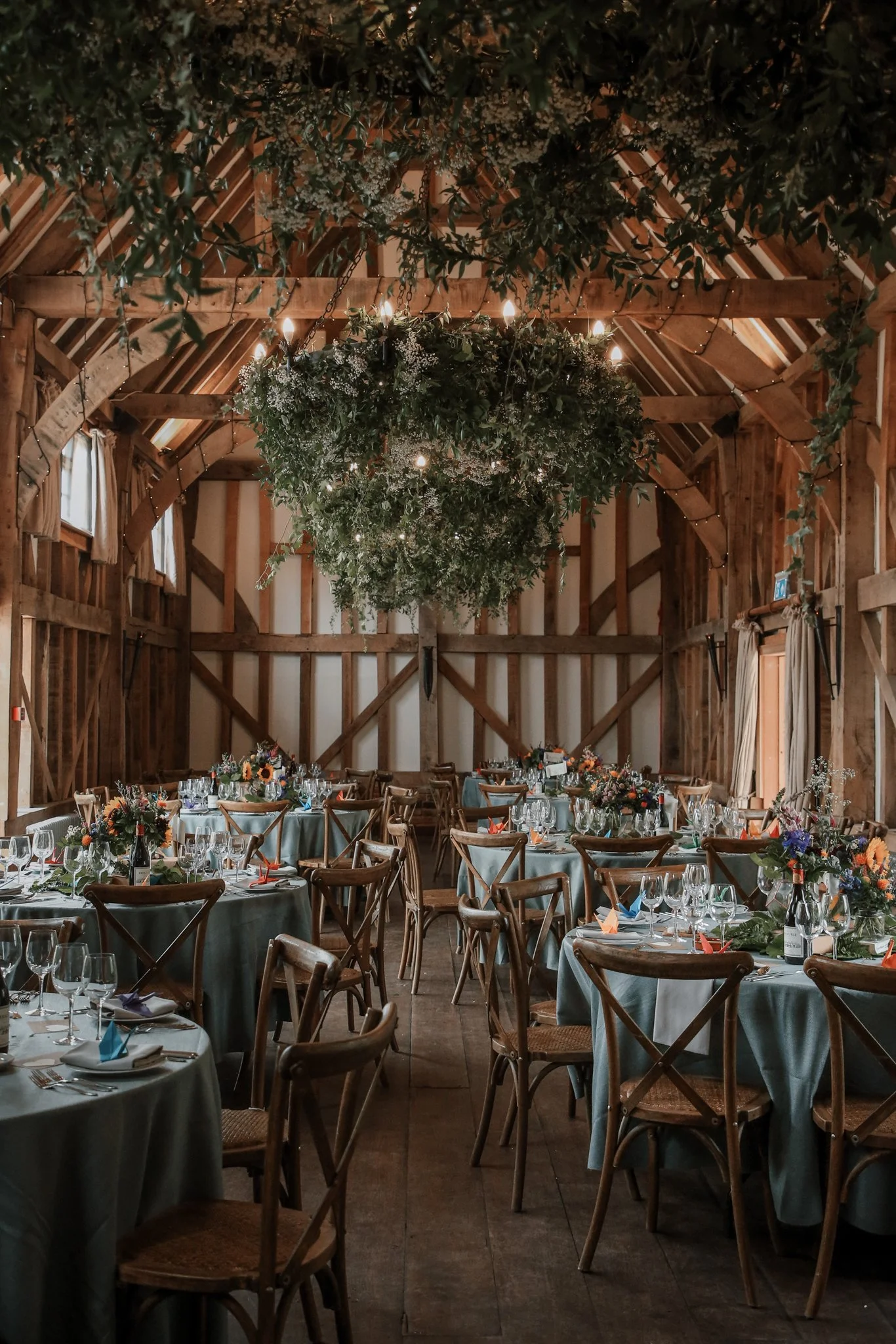 Elegant rustic wedding reception with tables covered in turquoise tablecloths, decorated with colorful flower arrangements, set for a formal dinner. Hanging floral chandelier and greenery adornments hang from the wooden ceiling of a barn-style venue 