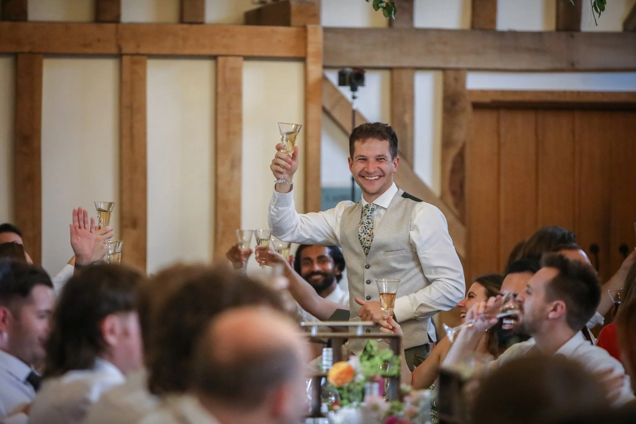 A man in a beige vest and floral tie smiles and holds up a glass of champagne during a celebration, surrounded by other people in a warmly decorated indoor setting.