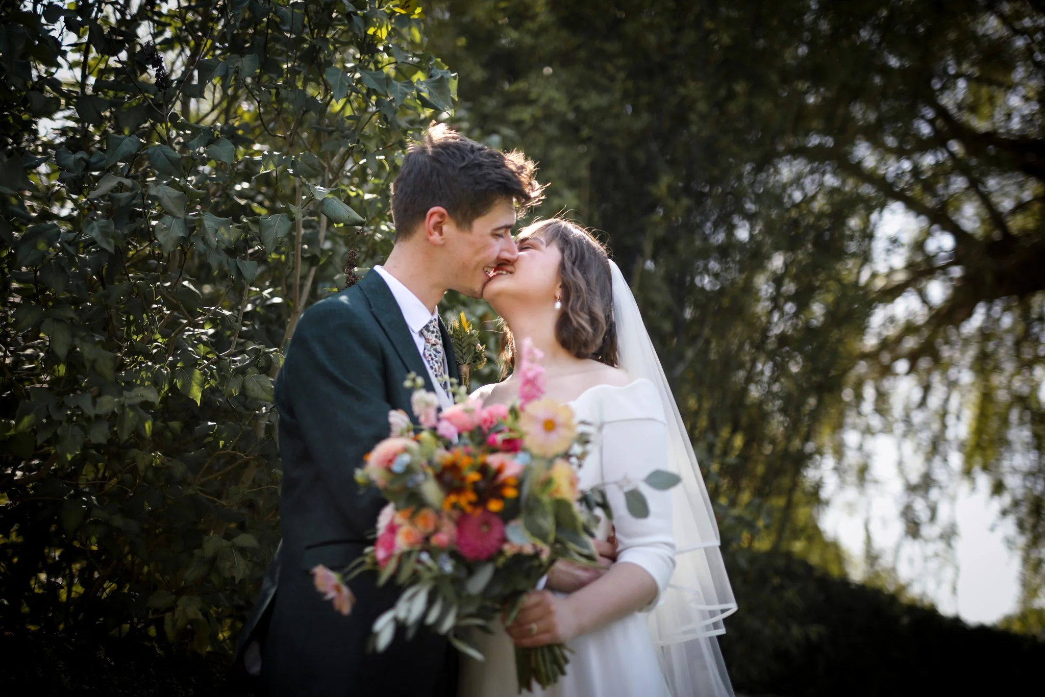 A bride and groom sharing a kiss outdoors, with the groom in a dark suit and a woman in a white dress holding a bouquet of pink, orange, and yellow flowers, surrounded by green trees.
