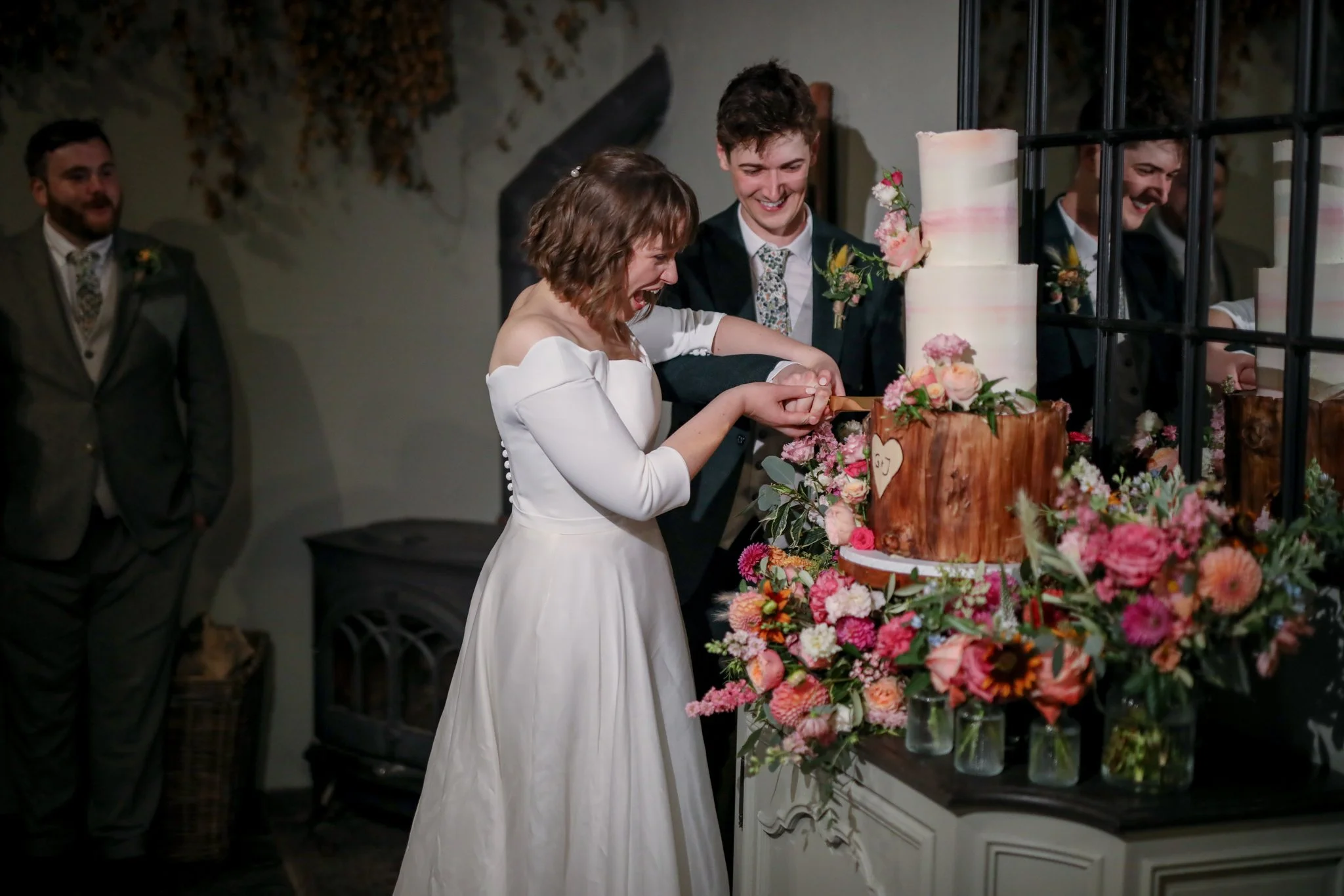 A woman and a man cutting a wedding cake decorated with pink and white flowers at a wedding reception, with guests in the background.