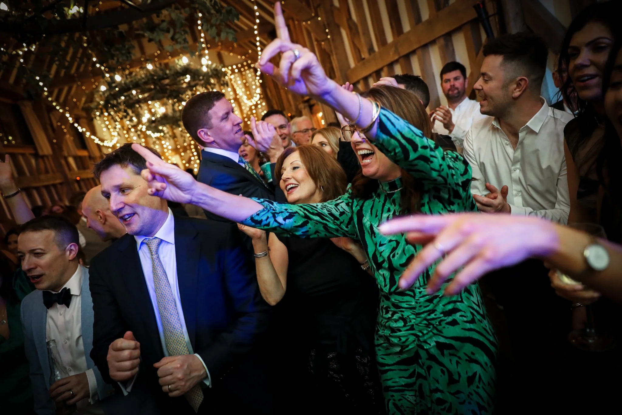 People dancing and celebrating at a festive event with wooden walls and string lights in the background.