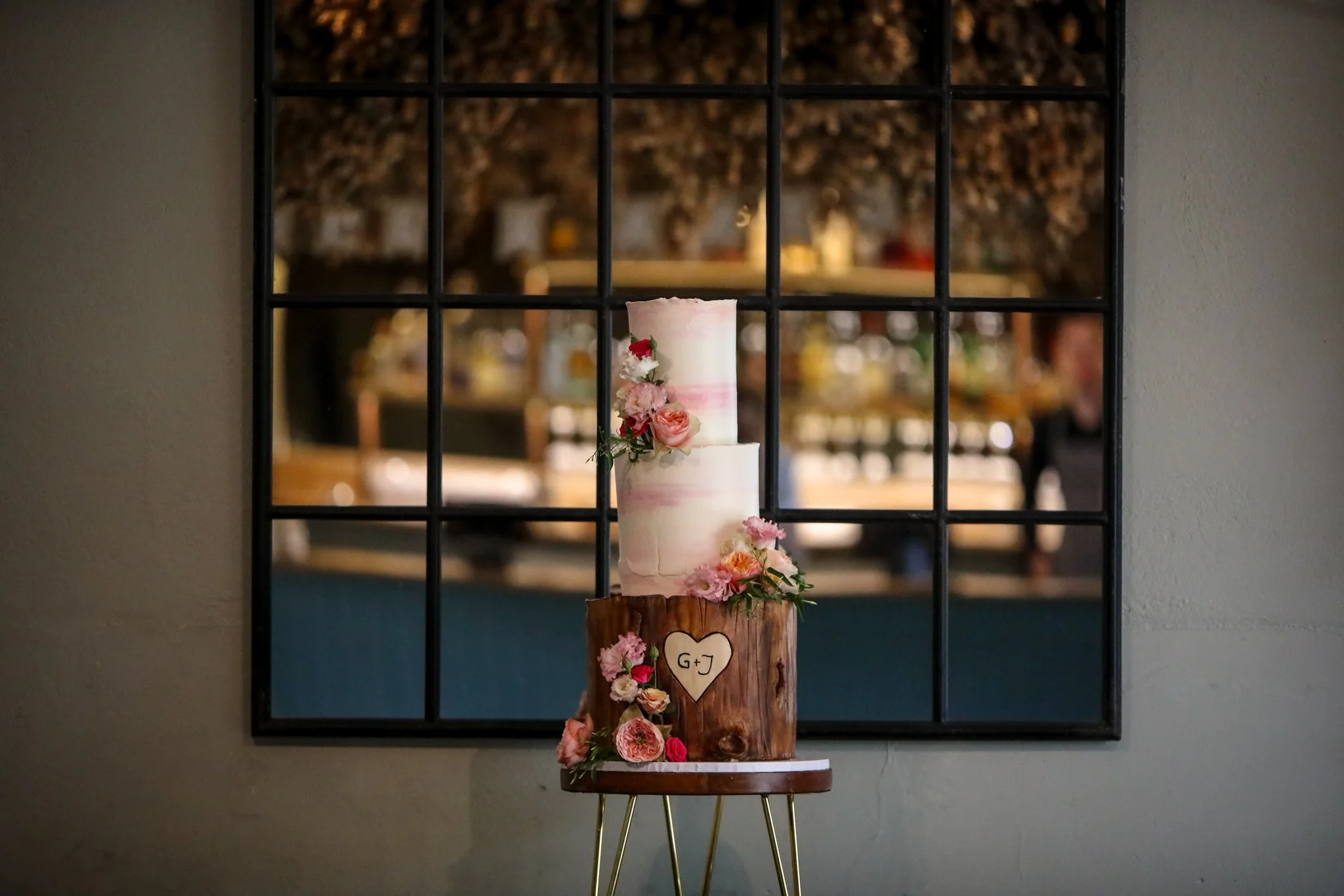 A tiered wedding cake decorated with pink and white flowers on a wooden stand against a wall with a windowpane mirror.