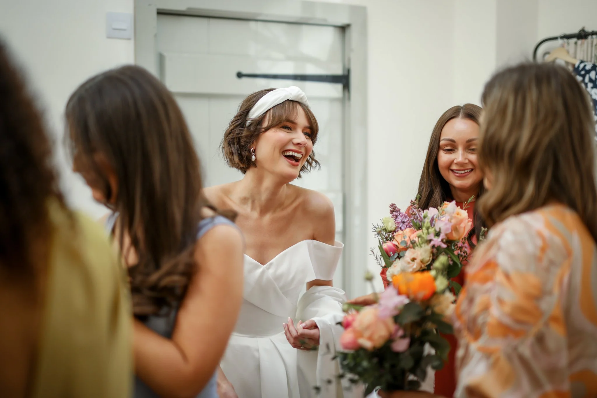 A woman in a white dress with a headband is smiling while holding a bouquet of flowers, surrounded by other women at a celebration or gathering.