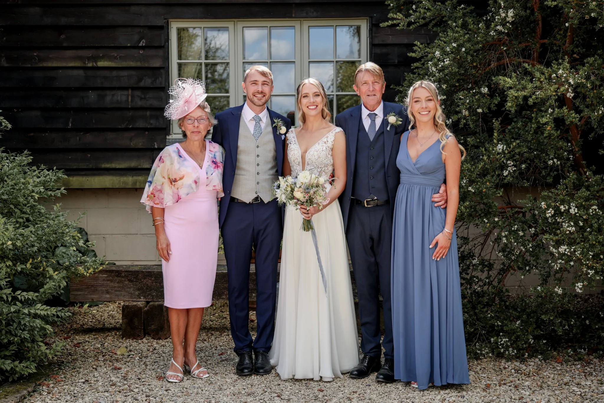 Group of six people, including a bride and groom, posing outdoors in front of a dark wooden building with a window, surrounded by greenery and white flowering bushes, on a wedding day.