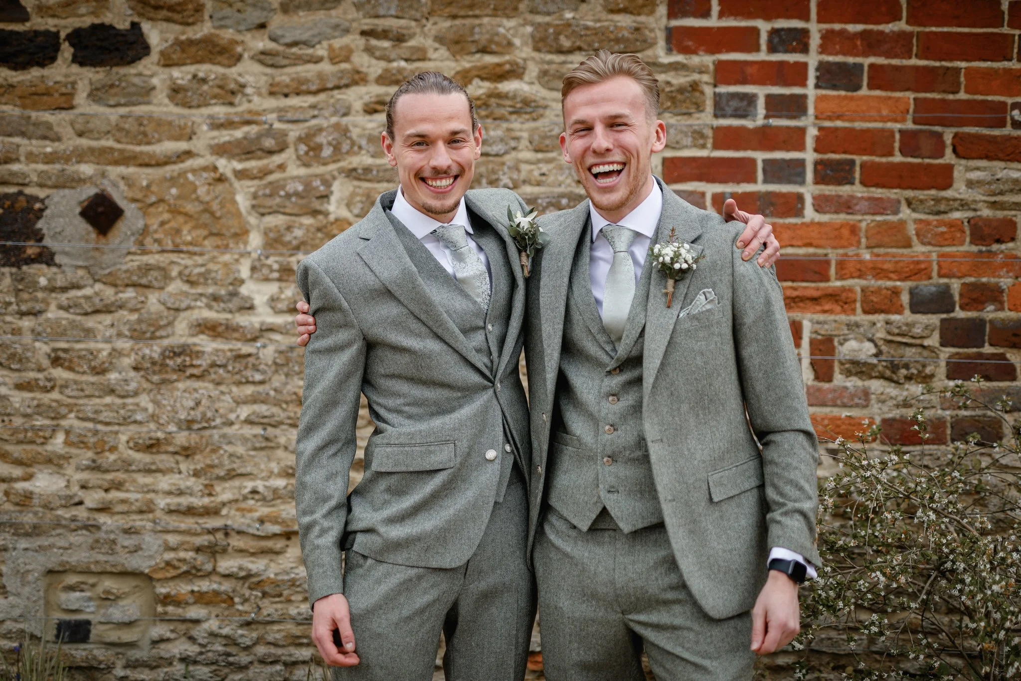 Two men in gray suits smiling and hugging each other outside in front of a brick wall.
