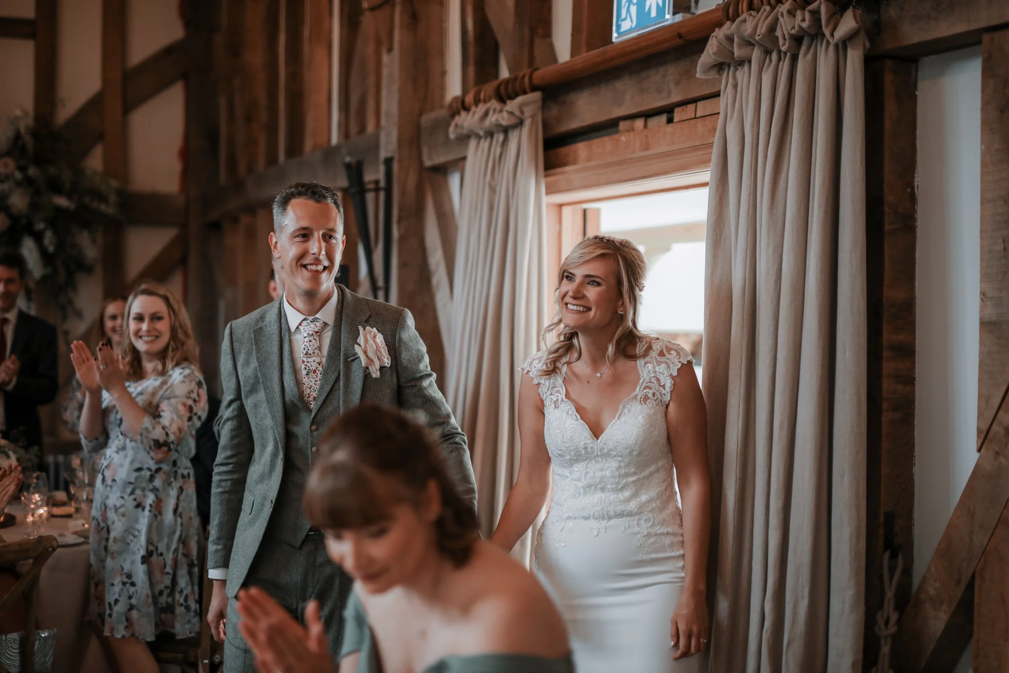 A bride and groom holding hands on their wedding day inside a rustic wooden venue. The groom is smiling and wearing a gray suit with a floral tie and a boutonniere, while the bride is smiling in a white lace wedding dress. Guests are seen clapping an