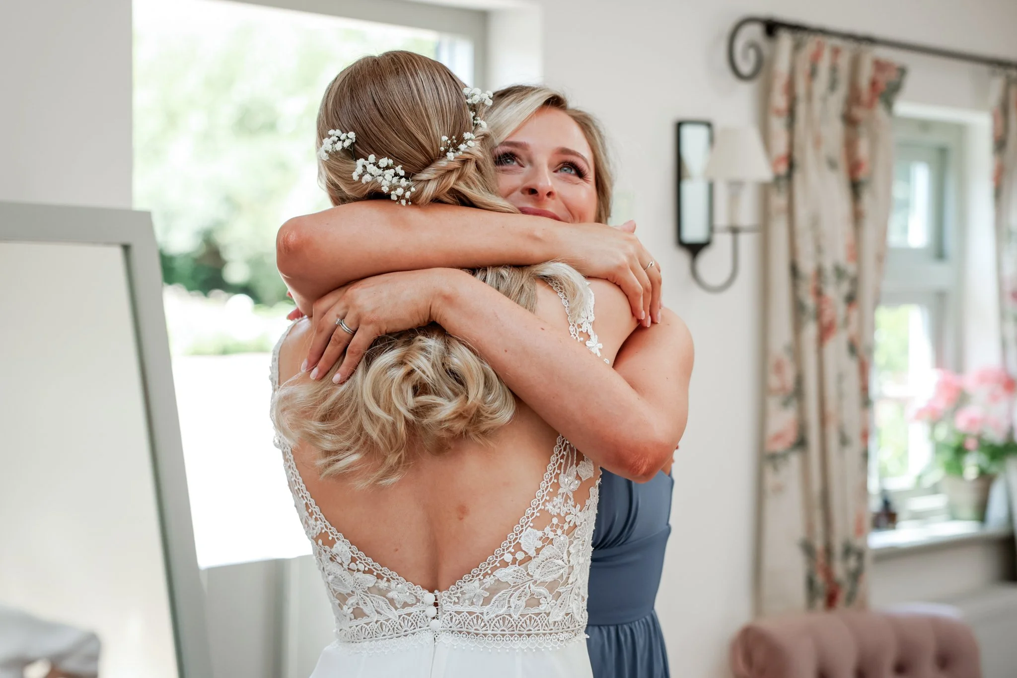 A bride hugging a woman, possibly her mother, inside a bright room with large windows and floral curtains. The bride has floral hair accessories and a lace wedding dress.