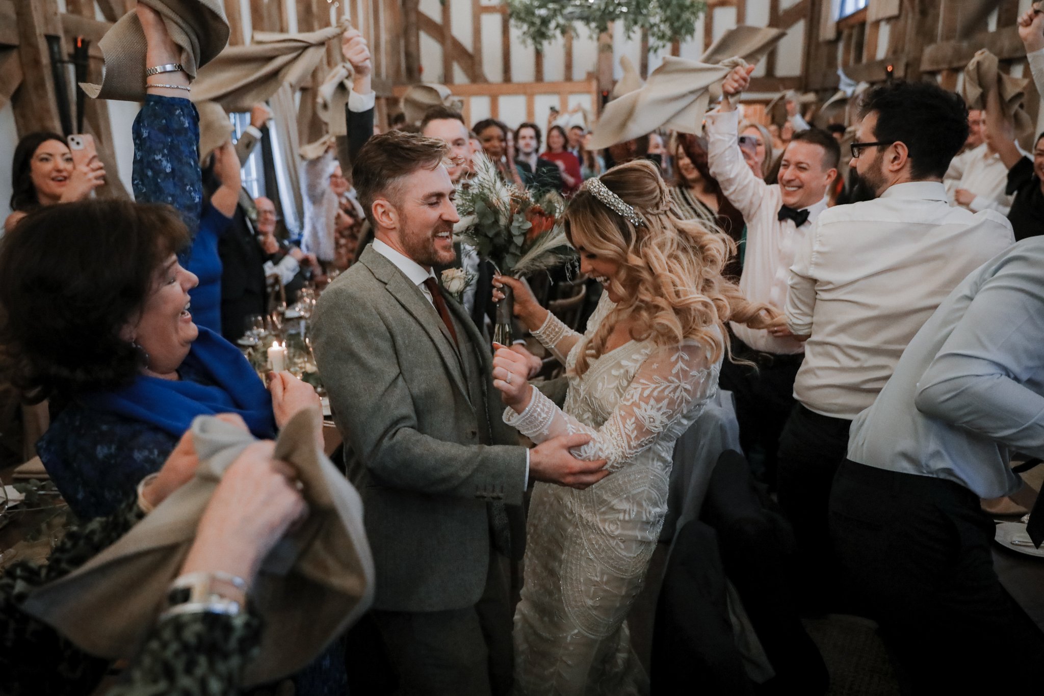 A bride and groom dancing at their wedding reception, surrounded by smiling guests in a rustic barn setting.