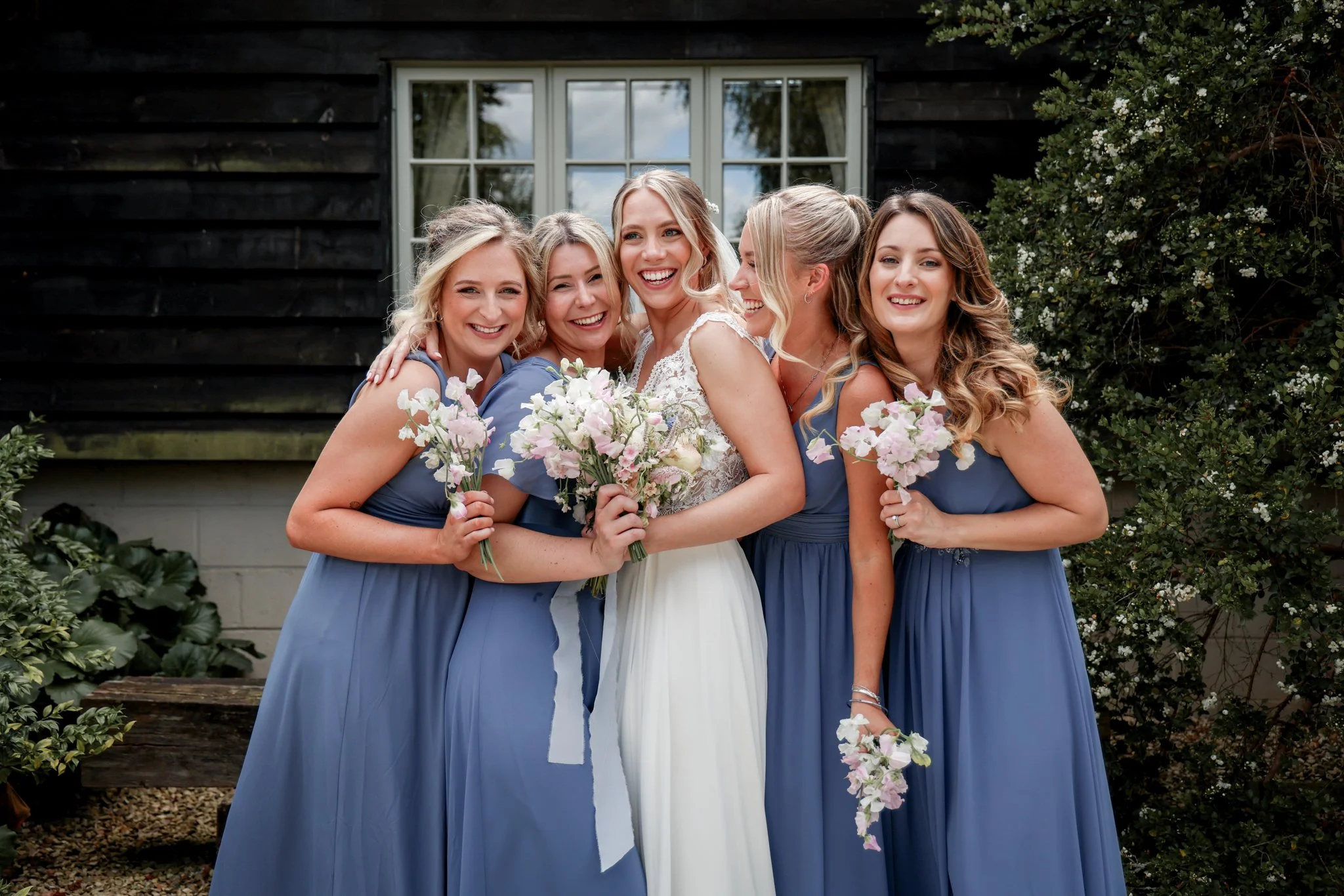 Group of six women smiling, dressed in formal wear, holding bouquets of flowers, outside near a black wooden building with a window and greenery.