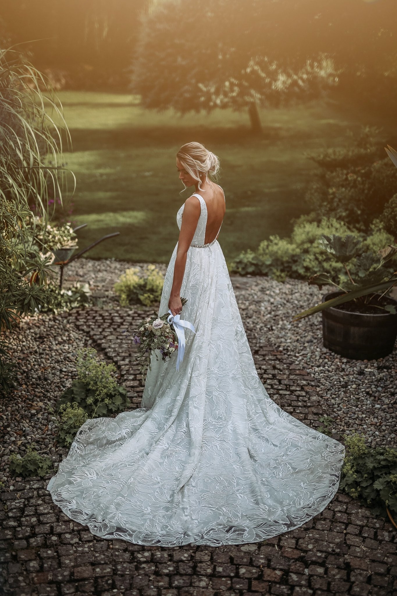 A woman in a white wedding dress holding a bouquet standing outdoors on a cobblestone path, surrounded by plants and trees with sunlight filtering through.