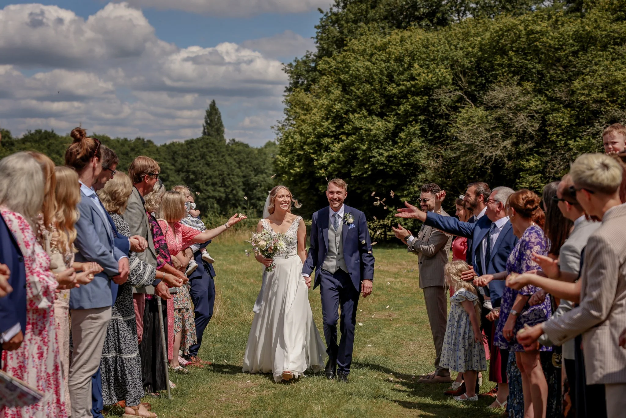 A bride and groom walking down the aisle during their outdoor wedding ceremony, surrounded by guests on both sides, with a large green tree and a partly cloudy sky in the background.