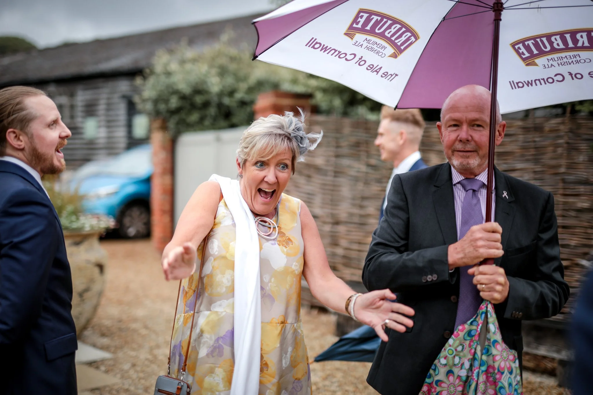 A group of people at an outdoor event, with one woman in a yellow floral dress and a fascinator, holding hands and smiling, and a man in a suit and purple tie holding an umbrella with the words 'tribe of Cornwall' on it, all in front of a brick and w