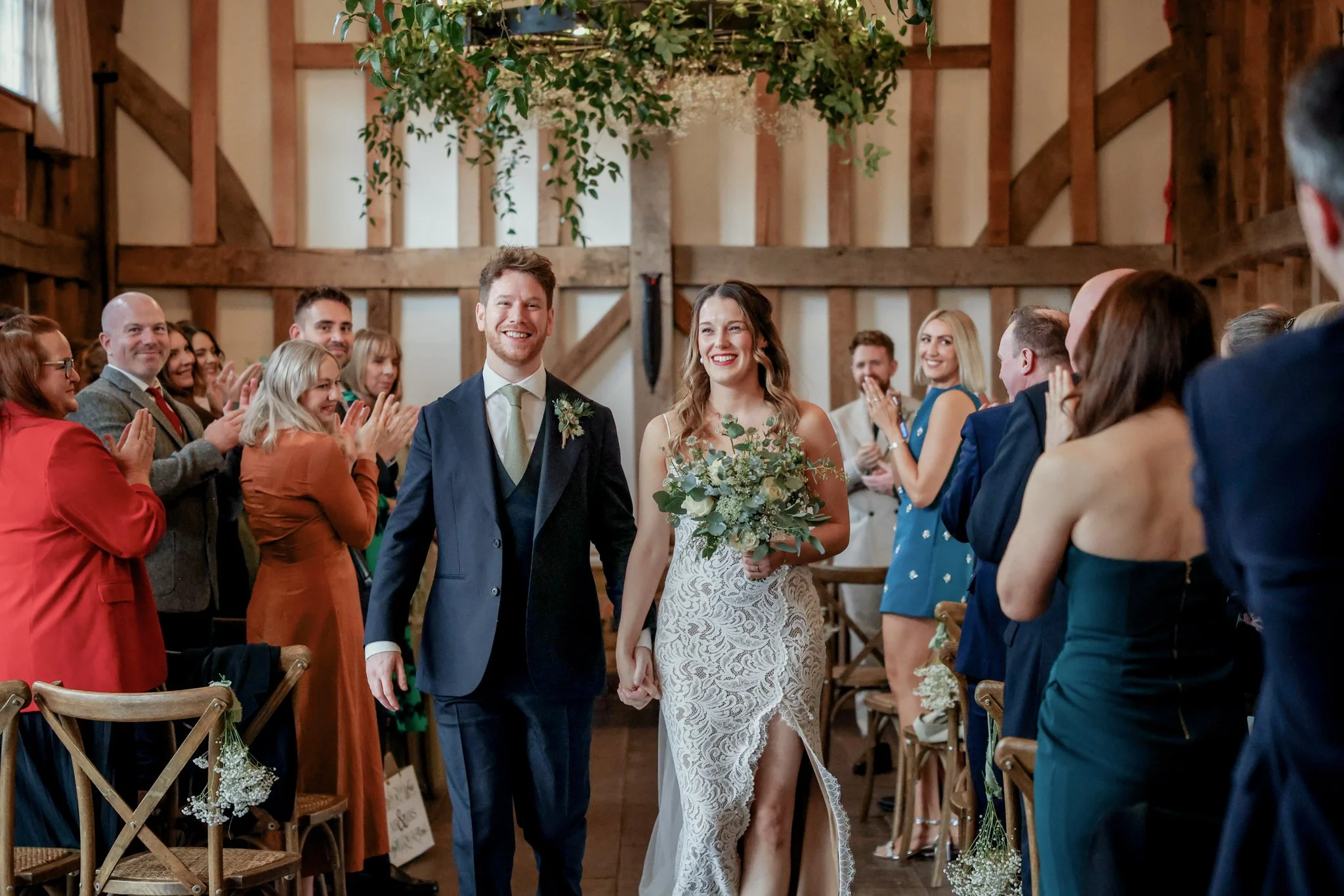 A bride and groom walking down the aisle hand-in-hand during their wedding ceremony in a rustic barn with wooden beams and greenery hanging from the ceiling, surrounded by smiling guests clapping and celebrating.